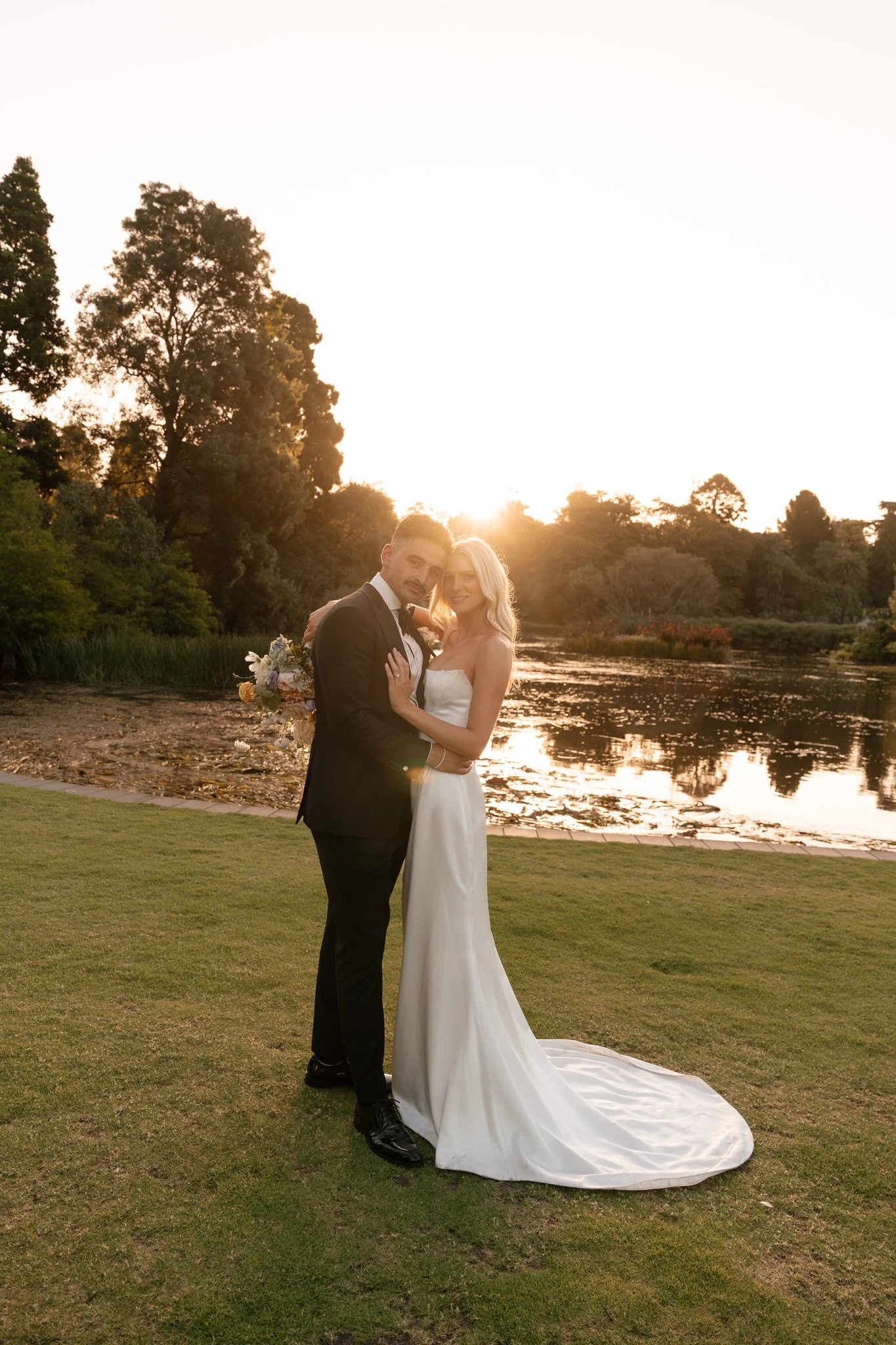 A bride and groom standing together outdoors at sunset by a pond, with trees in the background. The bride wears a white strapless gown and the groom wears a black tuxedo.