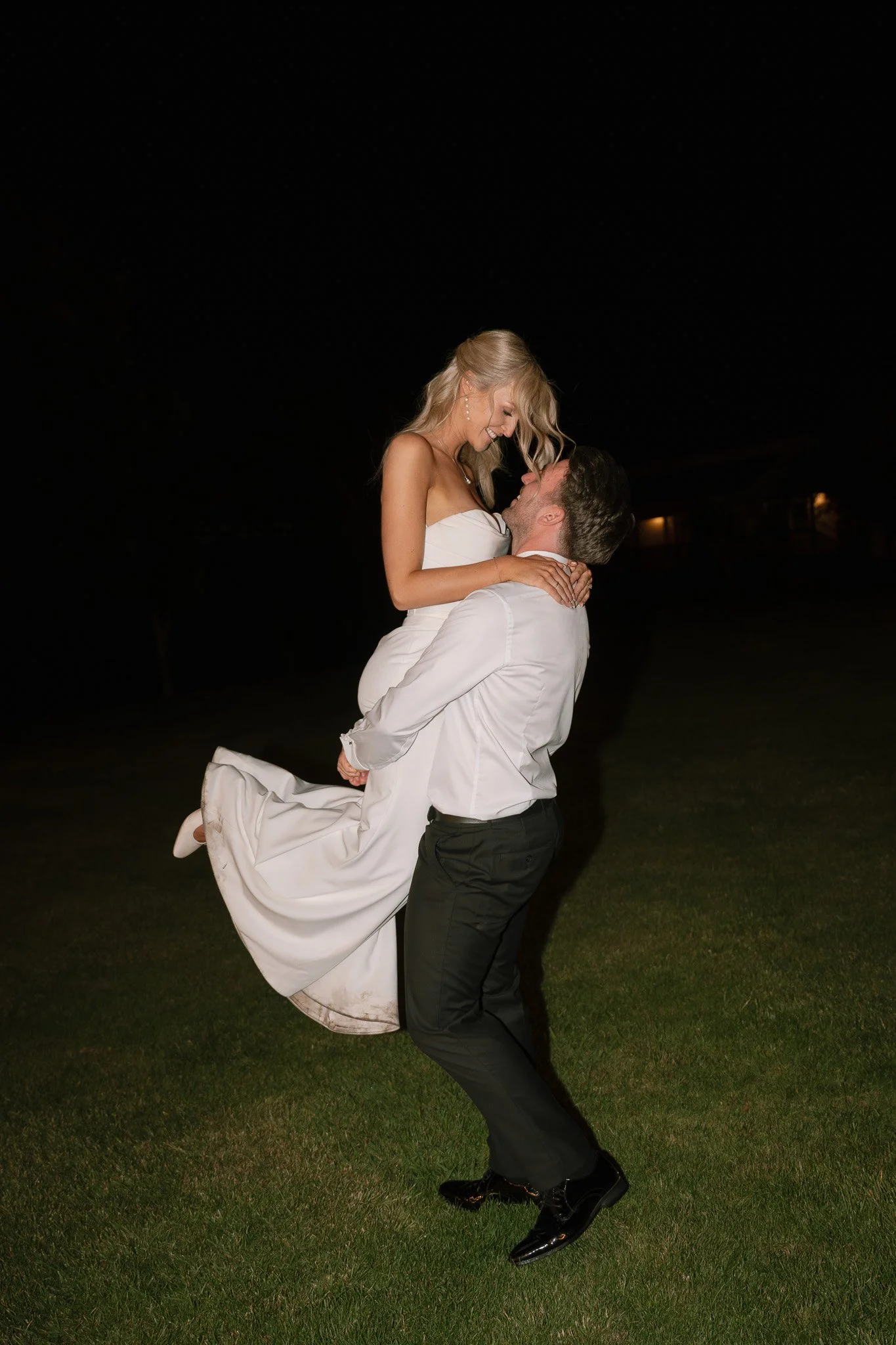 A man in black pants and a white shirt lifts a woman in a white dress into the air during nighttime outdoors. They are gazing at each other affectionately, with the woman smiling.