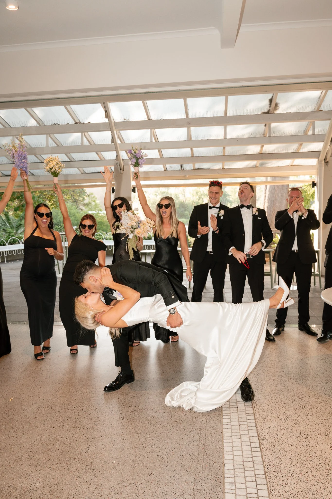 A newly married couple sharing a kiss while the bride's friends and groomsmen celebrate around them at a wedding reception.