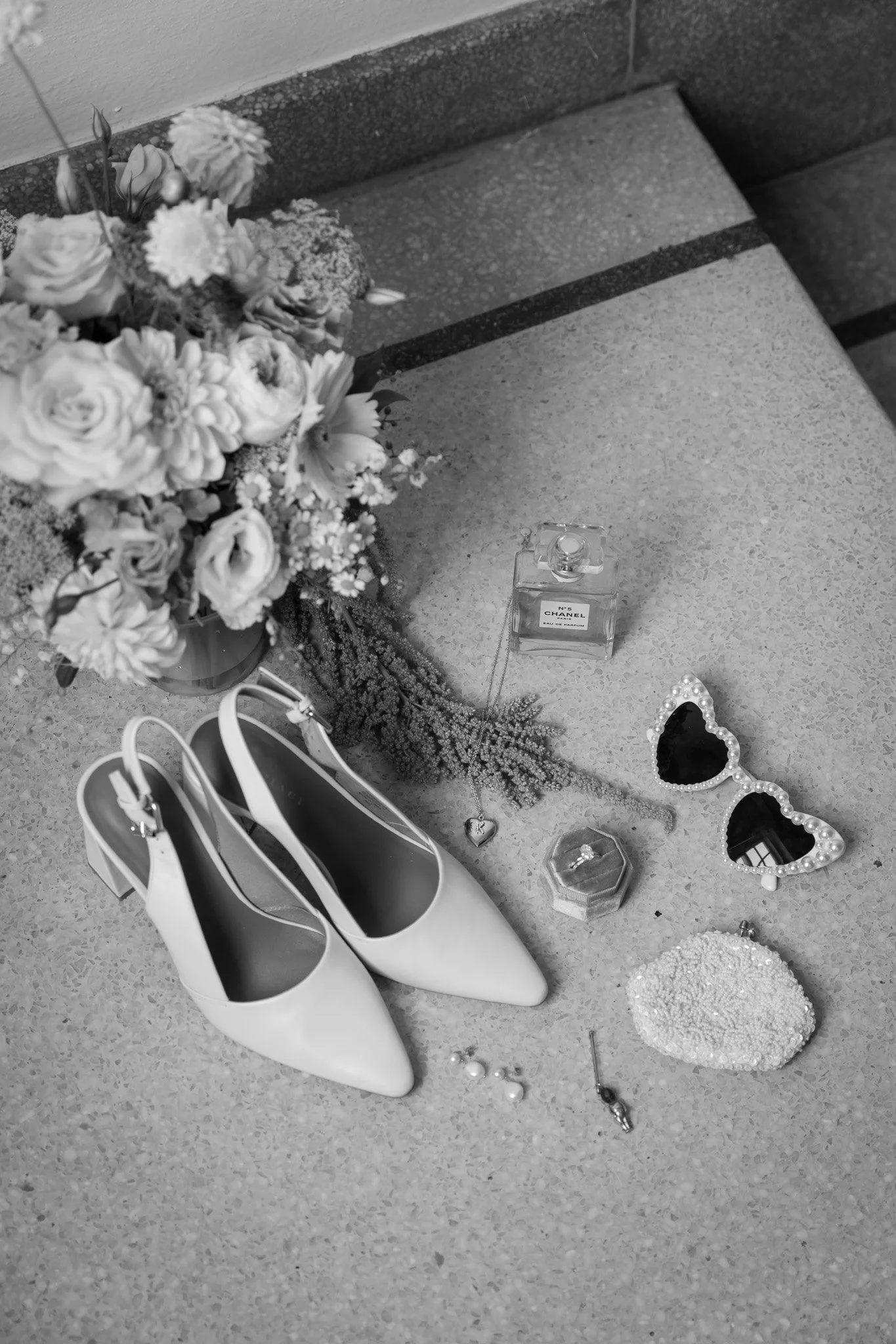 A collection of items on a concrete surface, including a bouquet of flowers, a pair of white high-heeled shoes, a bottle of Chanel perfume, a pair of heart-shaped sunglasses with pearl embellishments, a small ring box, a beaded clutch purse, two pear