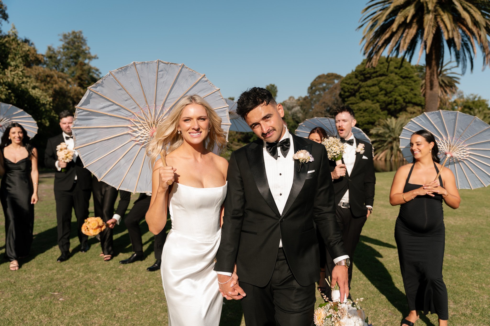 A newlywed couple walking in a park on a sunny day, holding hands, with bridesmaids and groomsmen in the background holding umbrellas.