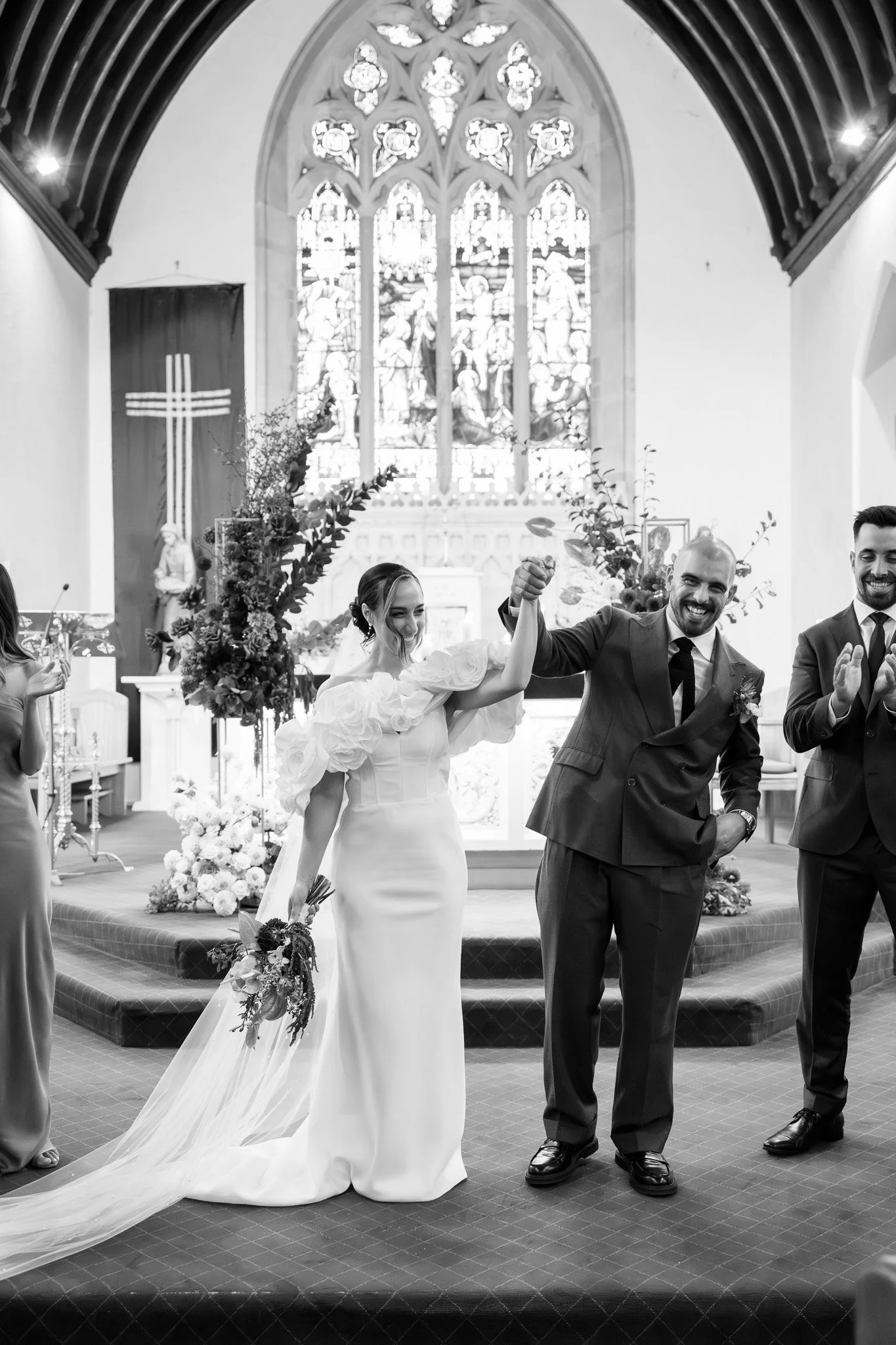 A bride and groom at their wedding ceremony inside a church, holding hands up in celebration, with bridesmaid and groomsman nearby, stained glass window, floral decorations, and church interior visible.