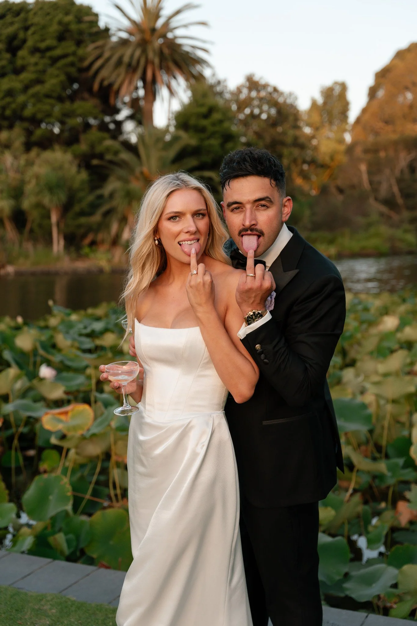 A bride and groom in wedding attire making playful gestures with their tongues out, standing outdoors near a pond with lily pads and lush trees.