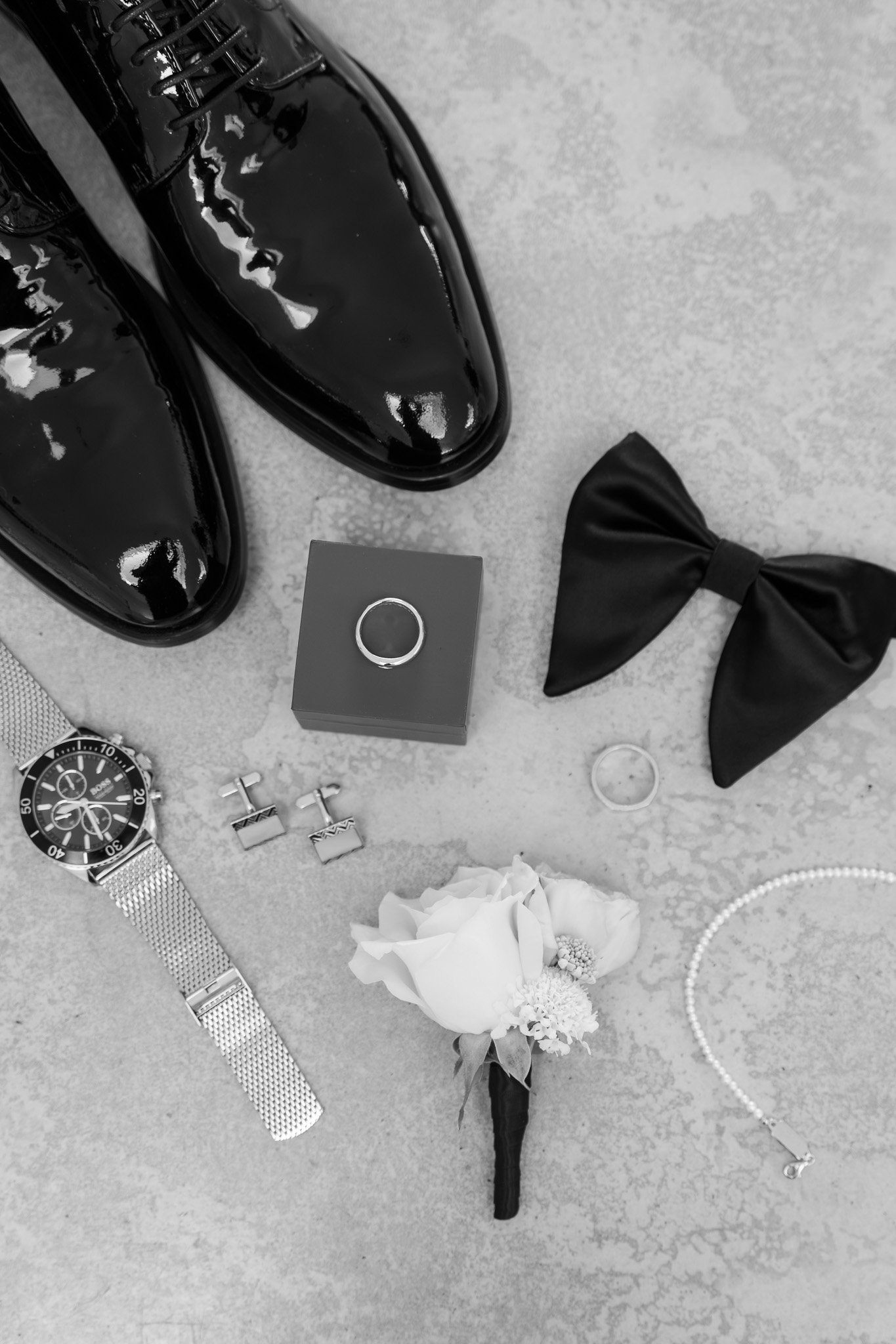 Black shiny dress shoes, a gray ring box with a ring, a black bow tie, a wristwatch with a silver band, a pair of cufflinks, a wedding band, a white flower boutonniere, a pearl bracelet, and a pair of earrings laid out on a flat surface.