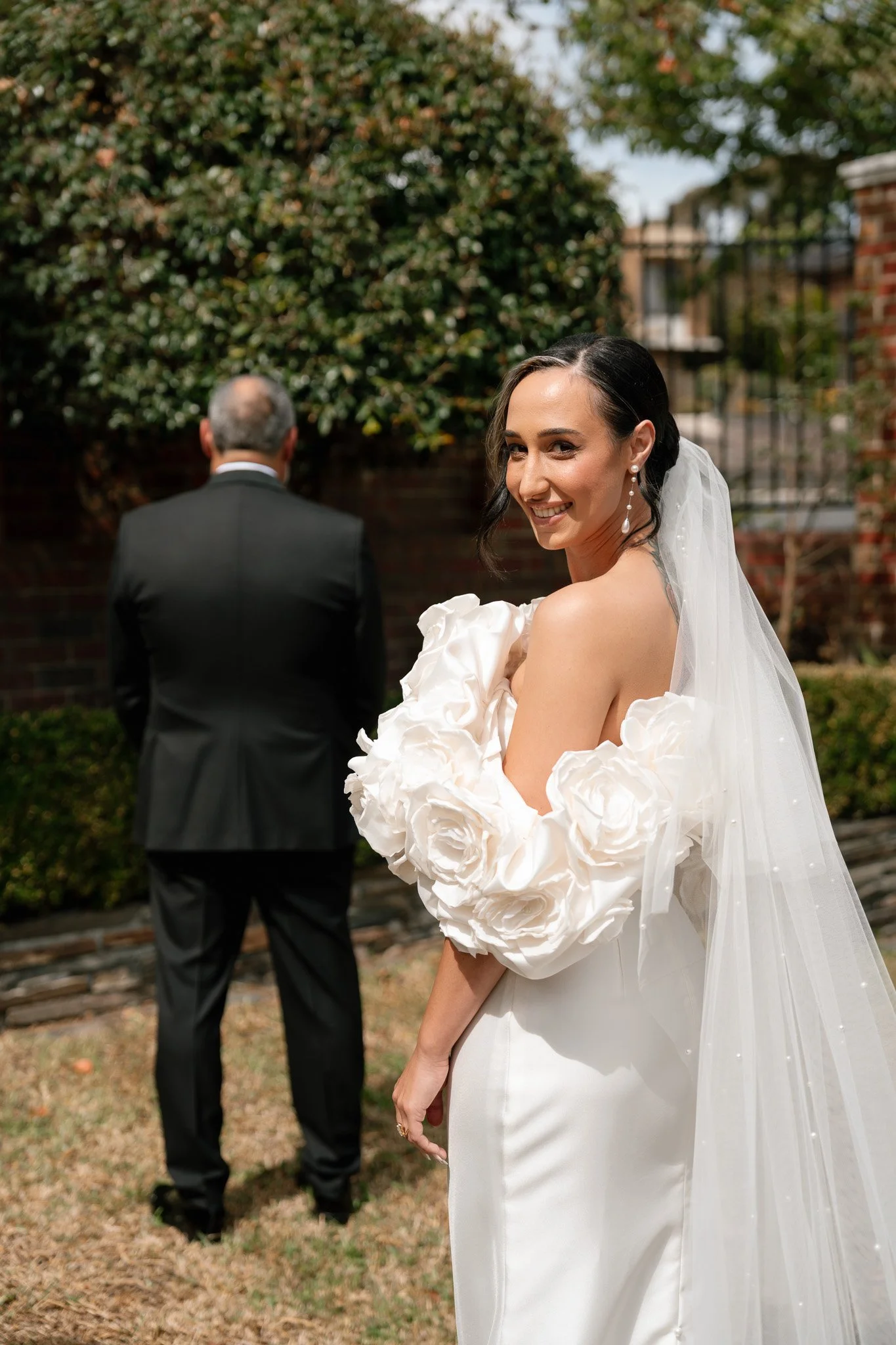 A smiling bride in a white wedding dress with large floral details on the sleeves, standing outdoors with a veil, and an older man in a black suit facing away in the background.