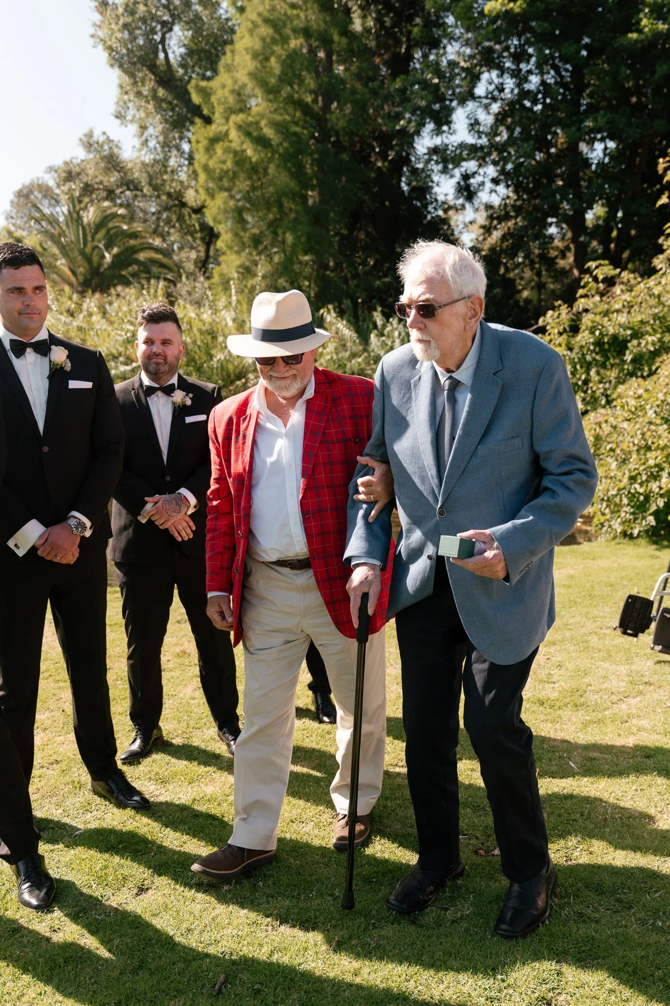 A group of men in formal attire standing outdoors in a garden with trees, during a wedding or special event. One elderly man with white hair and sunglasses is being assisted by another man with a cane.