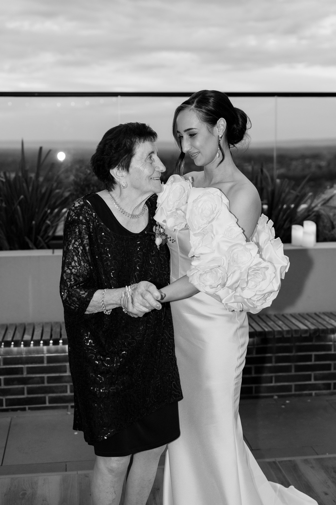 A bride and an older woman, likely her mother, smiling and holding hands during a wedding celebration on a rooftop balcony with a scenic sunset background.