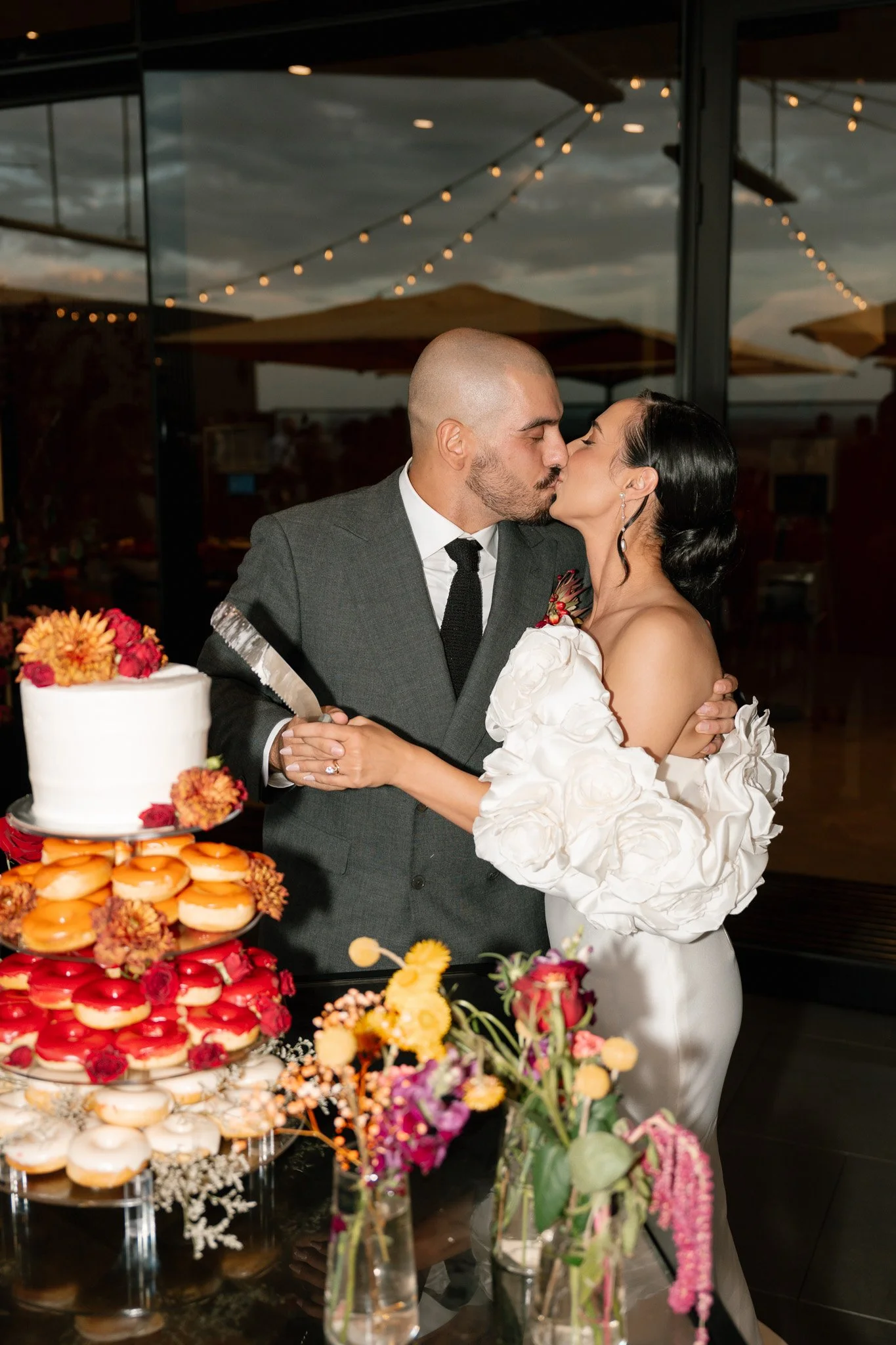 A newlywed couple sharing a kiss during their wedding reception. The groom is in a gray suit and the bride is in an off-the-shoulder white gown with floral details. A wedding cake and colorful flowers are on the table in front of them.