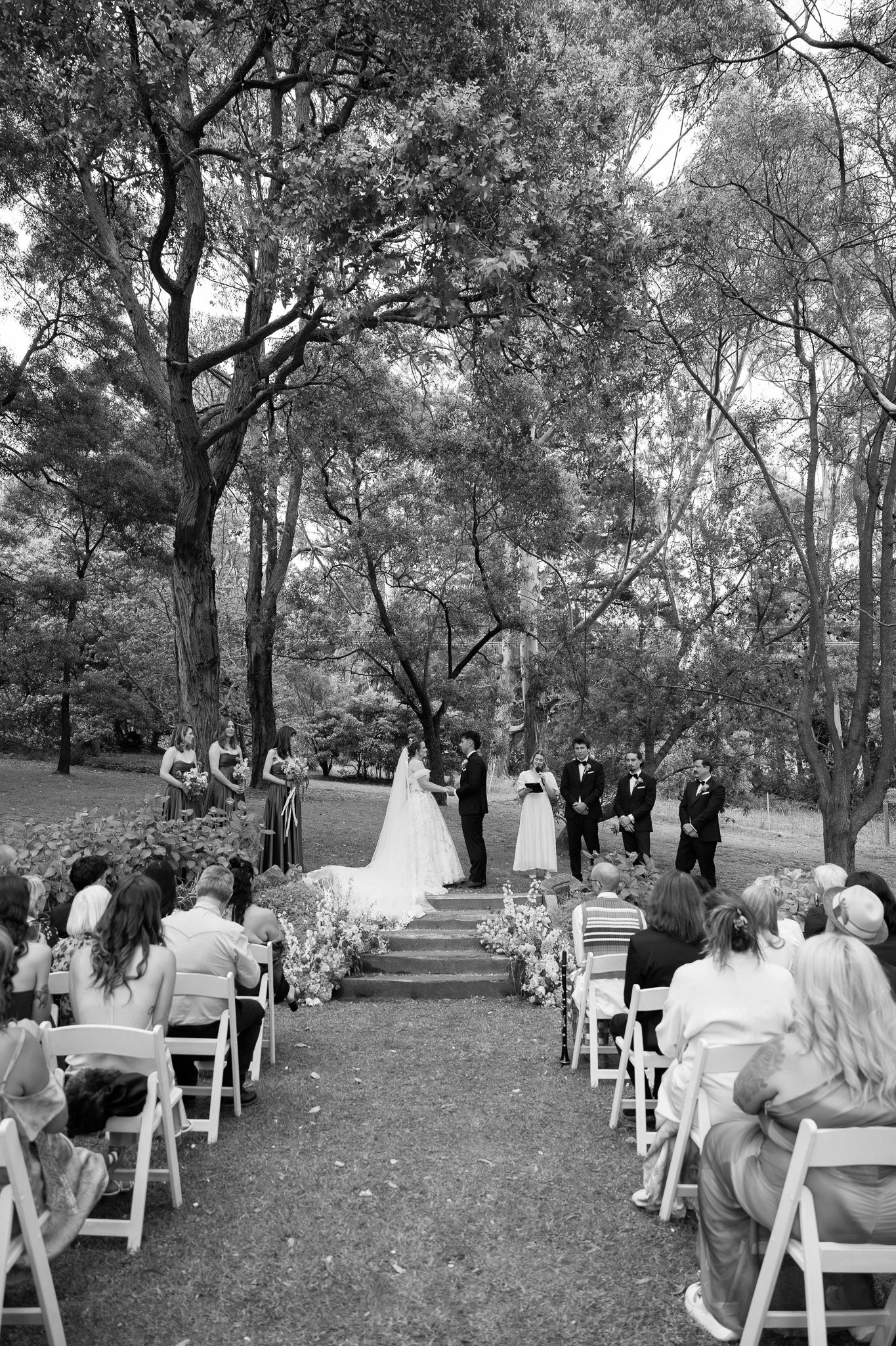 A black and white outdoor wedding ceremony with a bride and groom standing on a platform, surrounded by bridesmaids, groomsmen, and guests seated in white chairs under tall trees.
