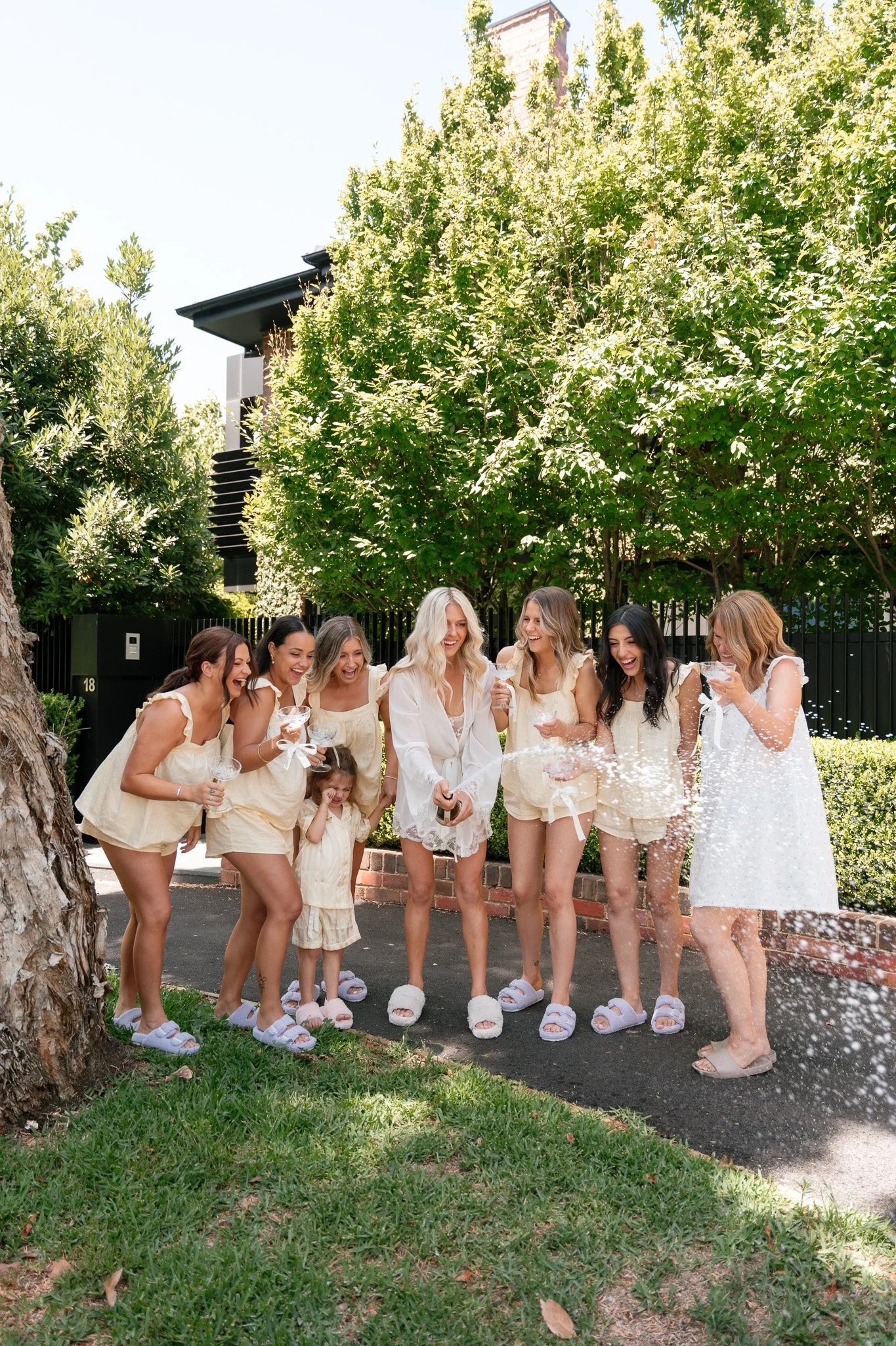 A group of women and a young girl celebrating outdoors, wearing light-colored clothing, and holding drinks while standing near a tree, with champagne being sprayed from a bottle.