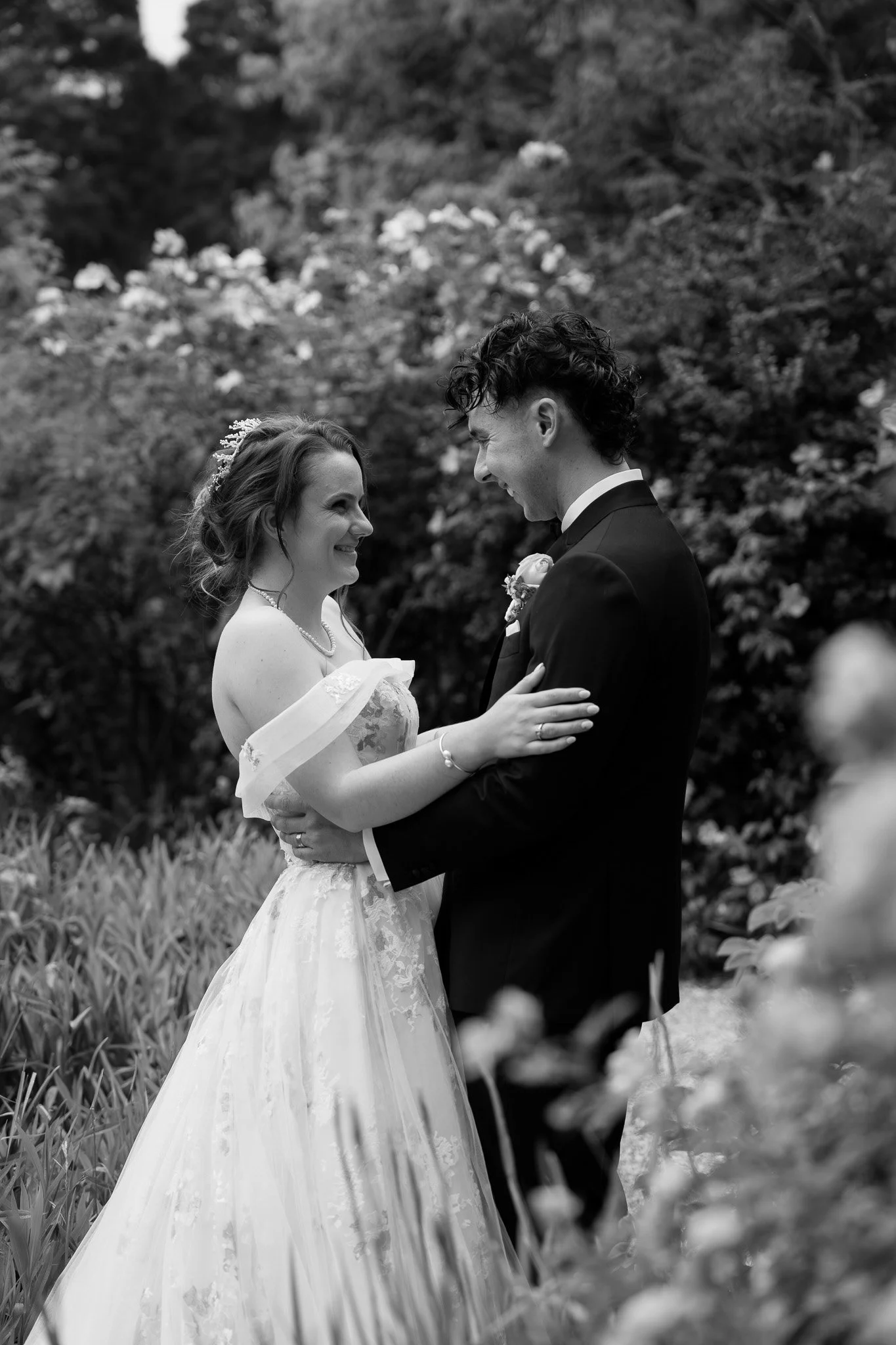 A bride and groom smiling and holding each other outdoors with trees and bushes in the background.