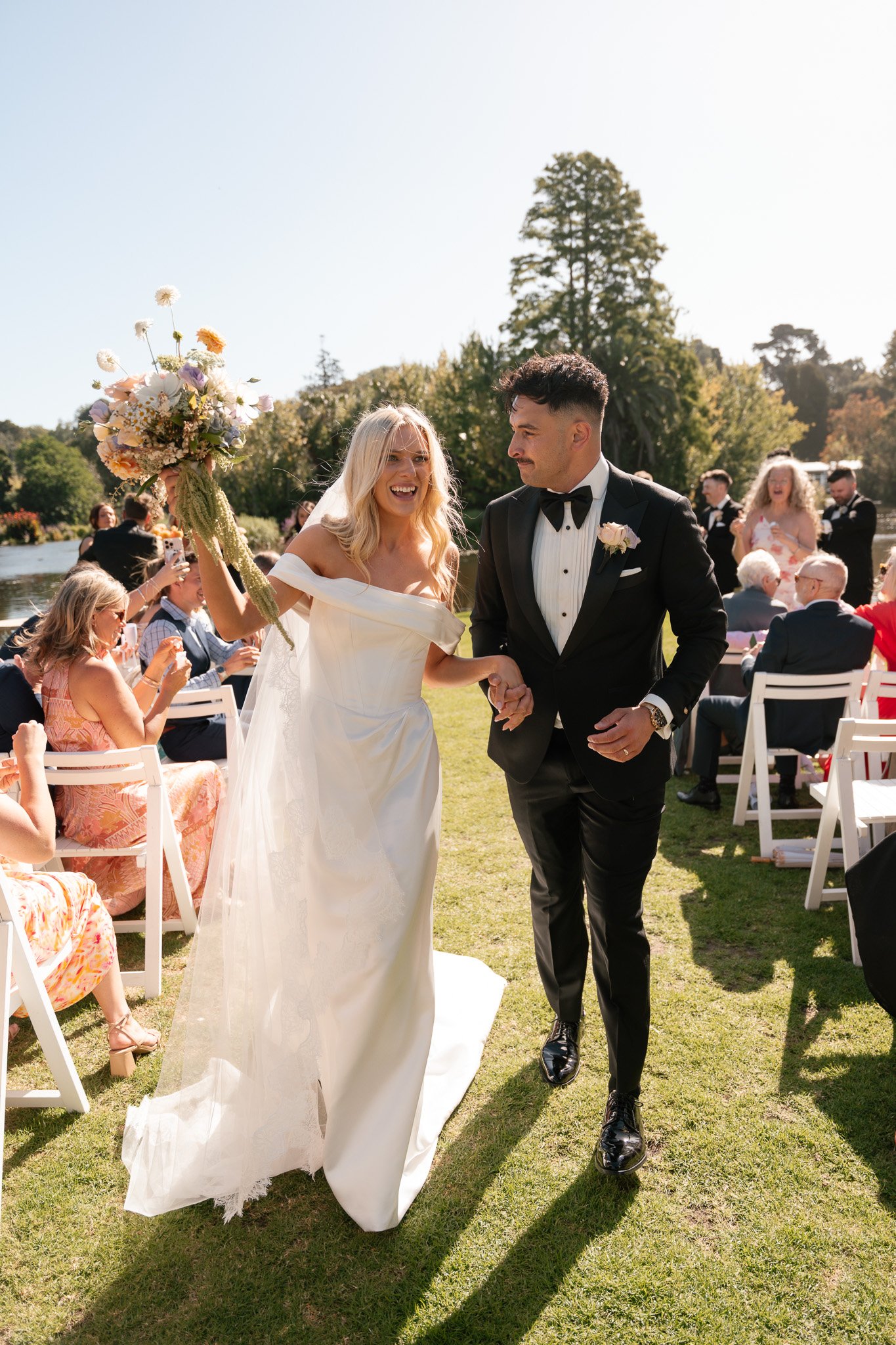 A newlywed couple walking outdoors during a wedding ceremony, with guests seated on white chairs on either side and a scenic green background.