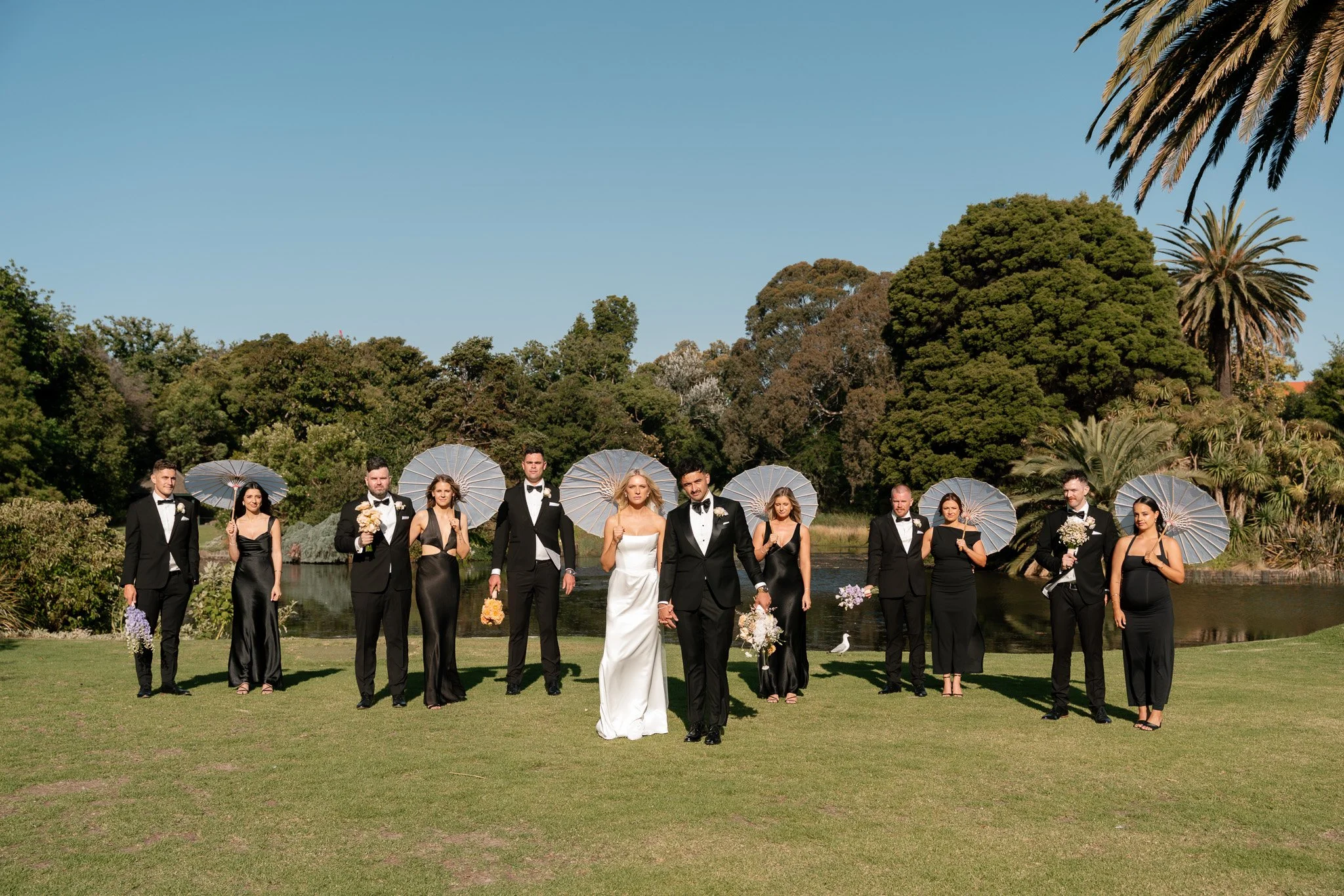 A wedding party of twelve people, dressed in black and white, standing outdoors on a grassy area near a pond with trees in the background. The bride and groom are in the center, holding hands, with bridesmaids and groomsmen surrounding them, some hol