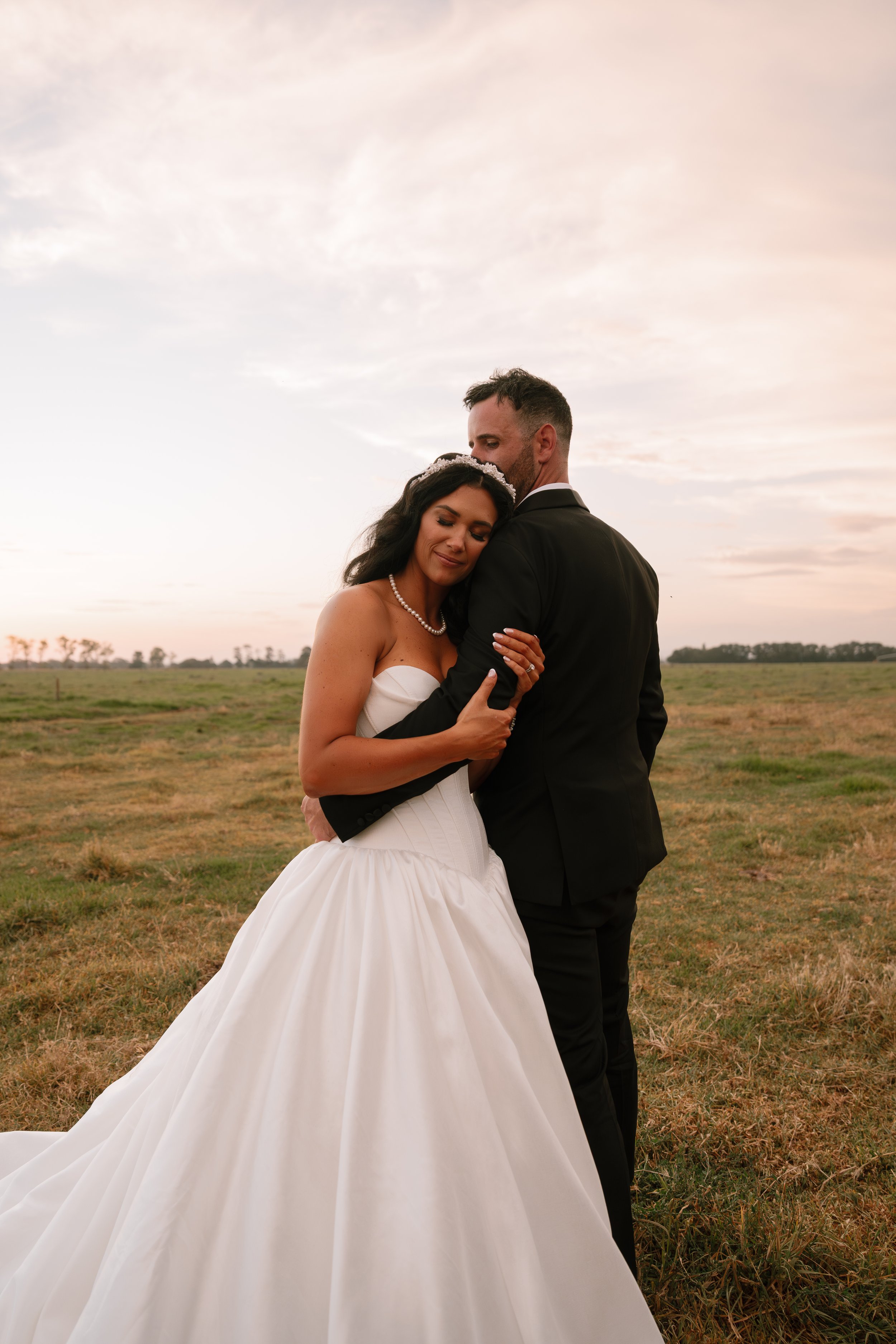 A bride and groom embrace outdoors during sunset. The bride wears a strapless white wedding gown with a full skirt and a pearl necklace, and the groom wears a black suit with a white shirt. They are standing in a grassy field with a cloudy sky in the background.