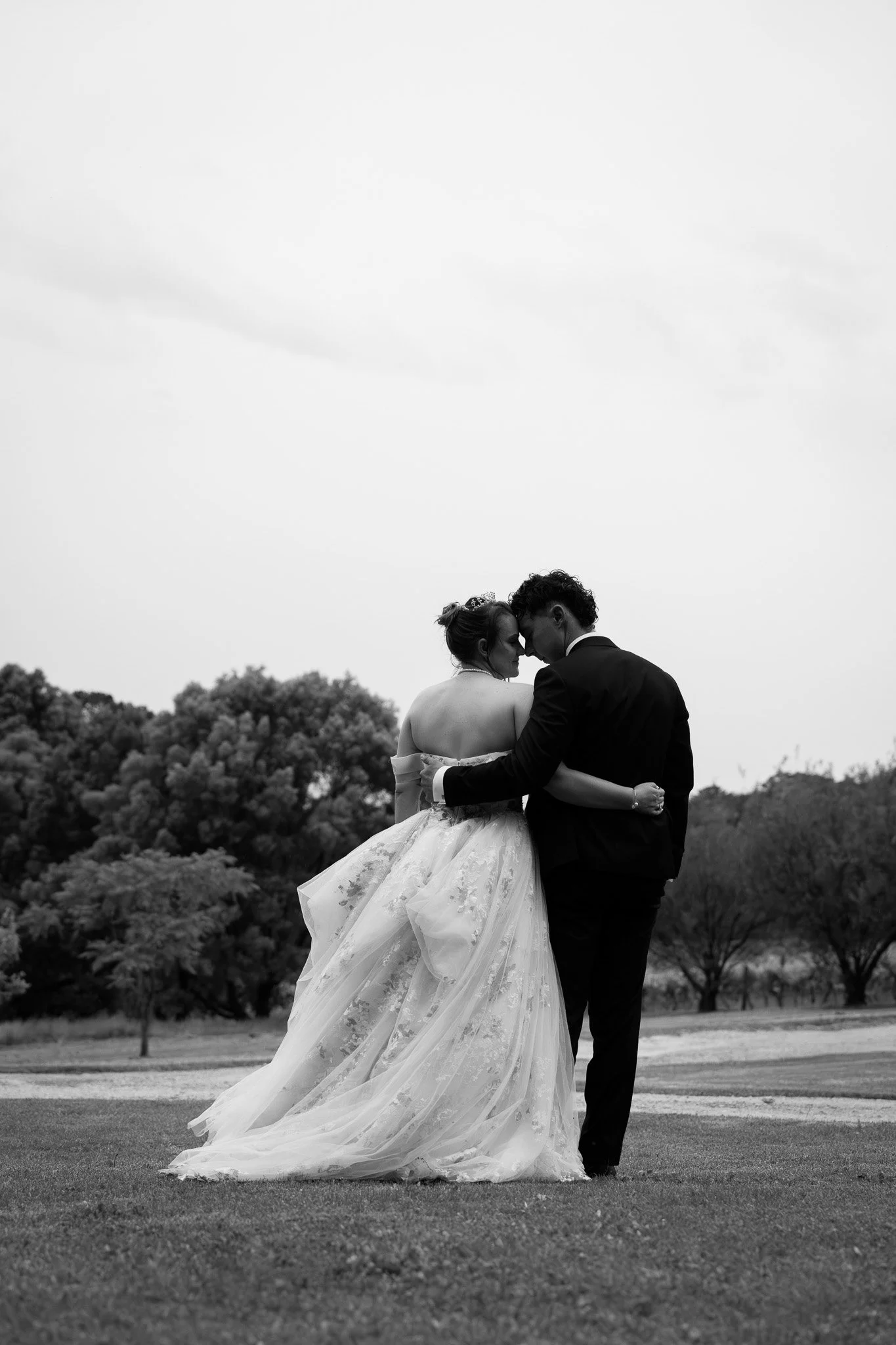 A black-and-white photo of a bride and groom embracing outdoors, with trees and an open sky in the background.