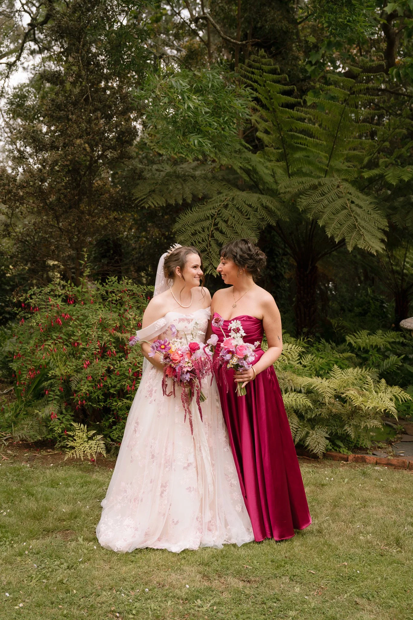 Two women wearing formal dresses, one in a white wedding gown with a veil and the other in a magenta dress, standing close together outdoors and smiling at each other while holding bouquets of flowers. There are green trees and plants in the backgrou