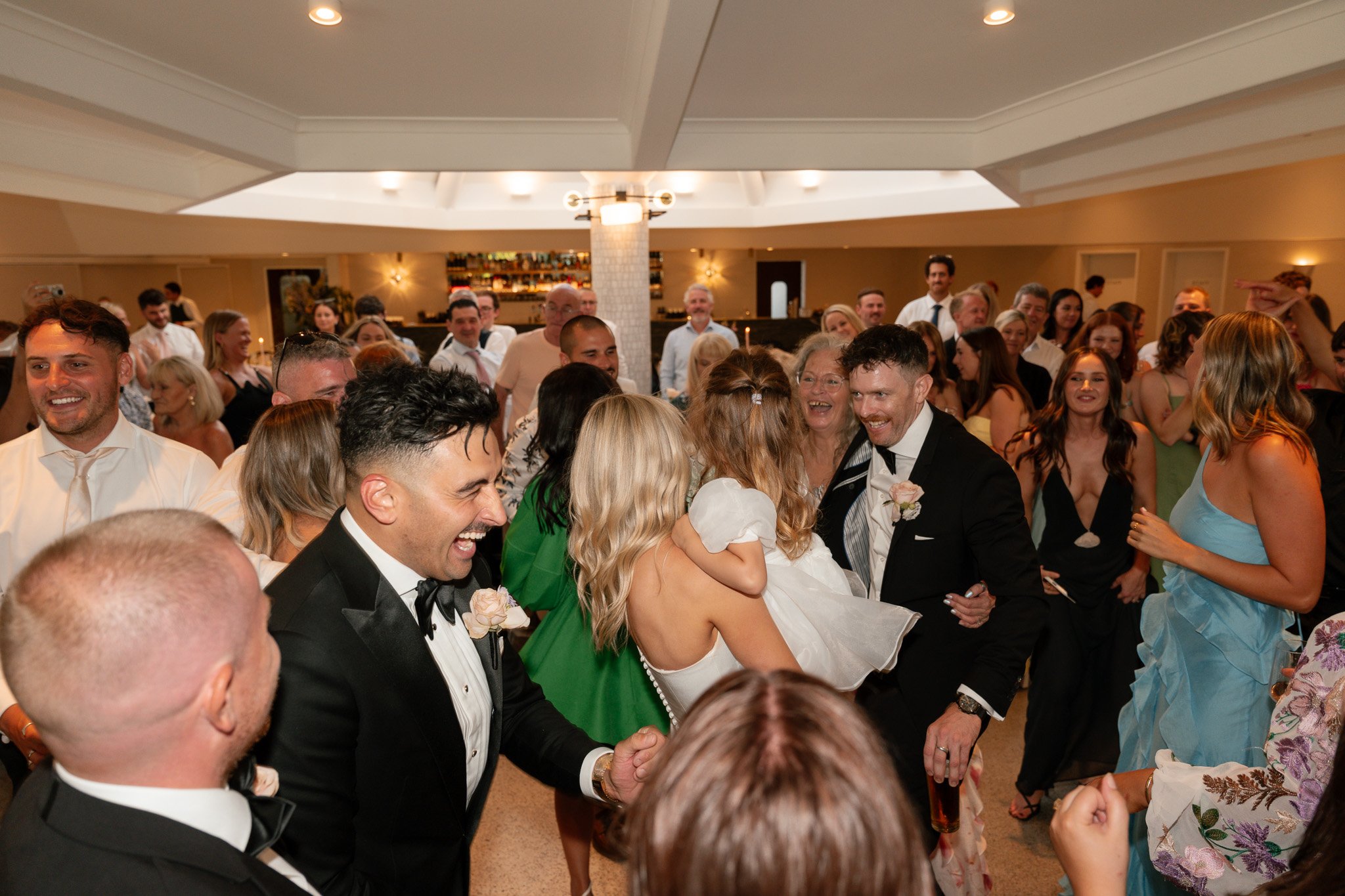 A lively wedding reception with many guests dancing and celebrating indoors, smiling and enjoying the moment.