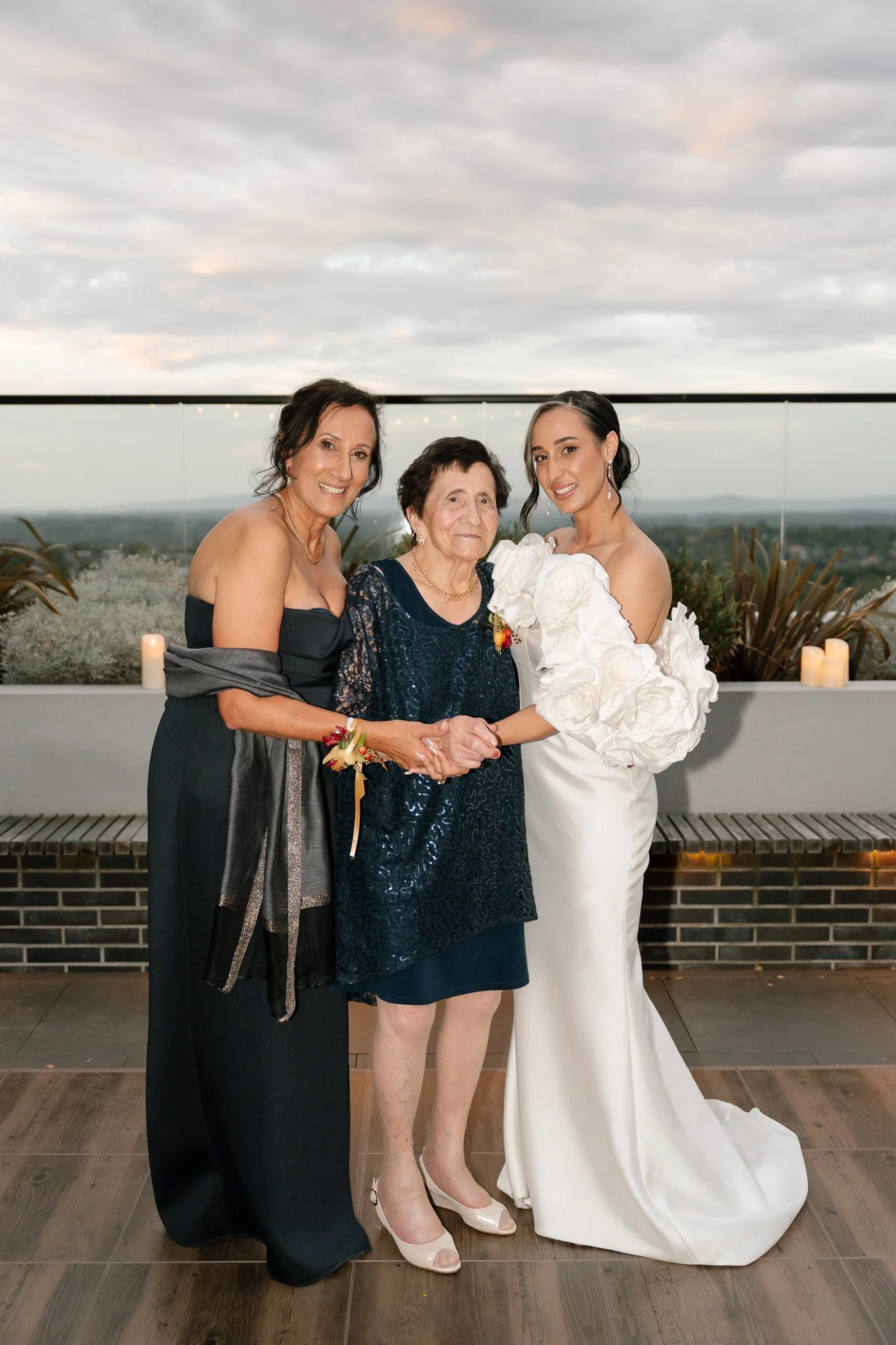 Three women holding hands and smiling at a wedding reception on a rooftop with a scenic city view and candles in the background.