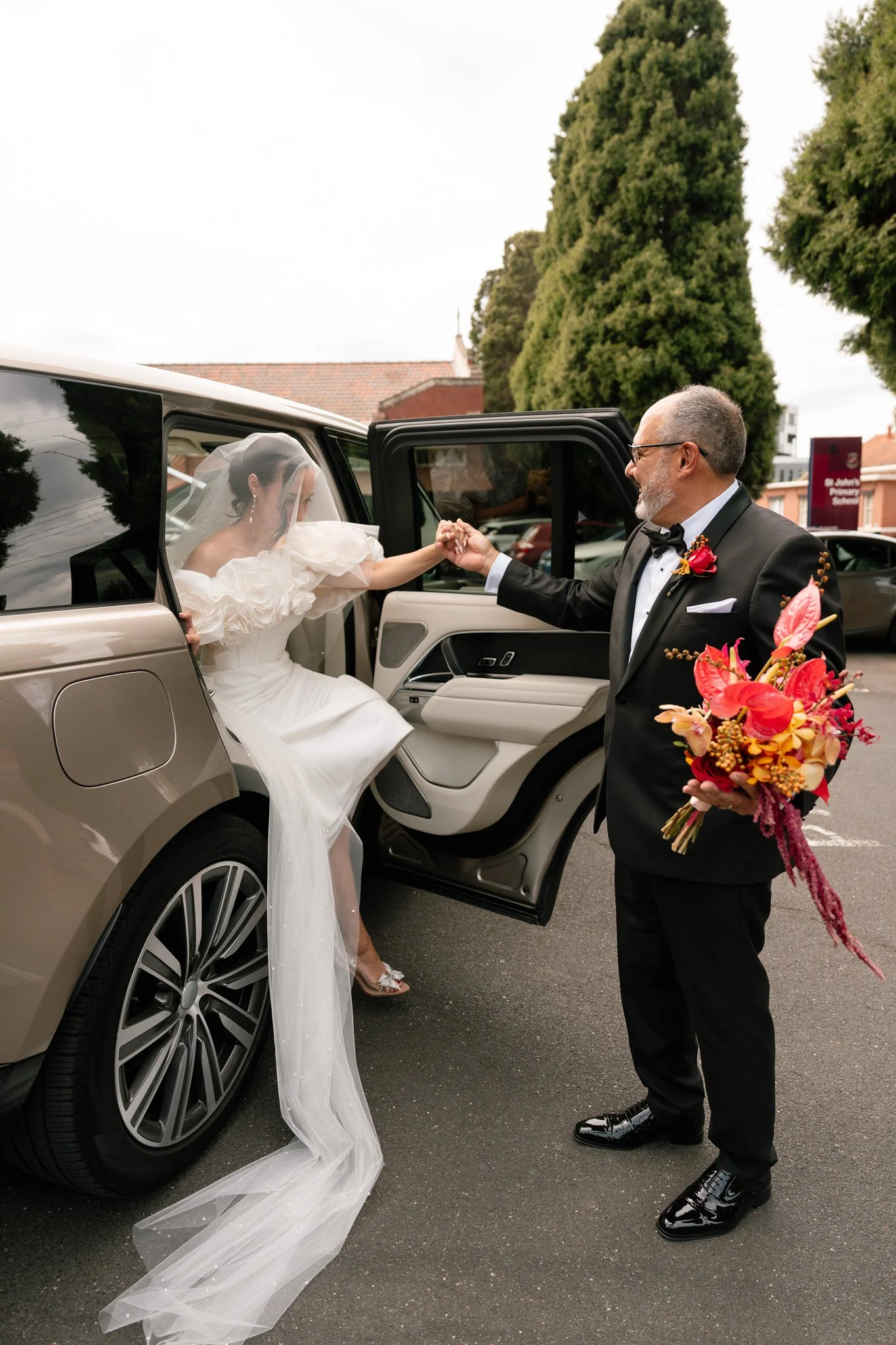 A bride in a white wedding dress and veil is exiting a car and shaking hands with a man in a tuxedo holding a bouquet of flowers.