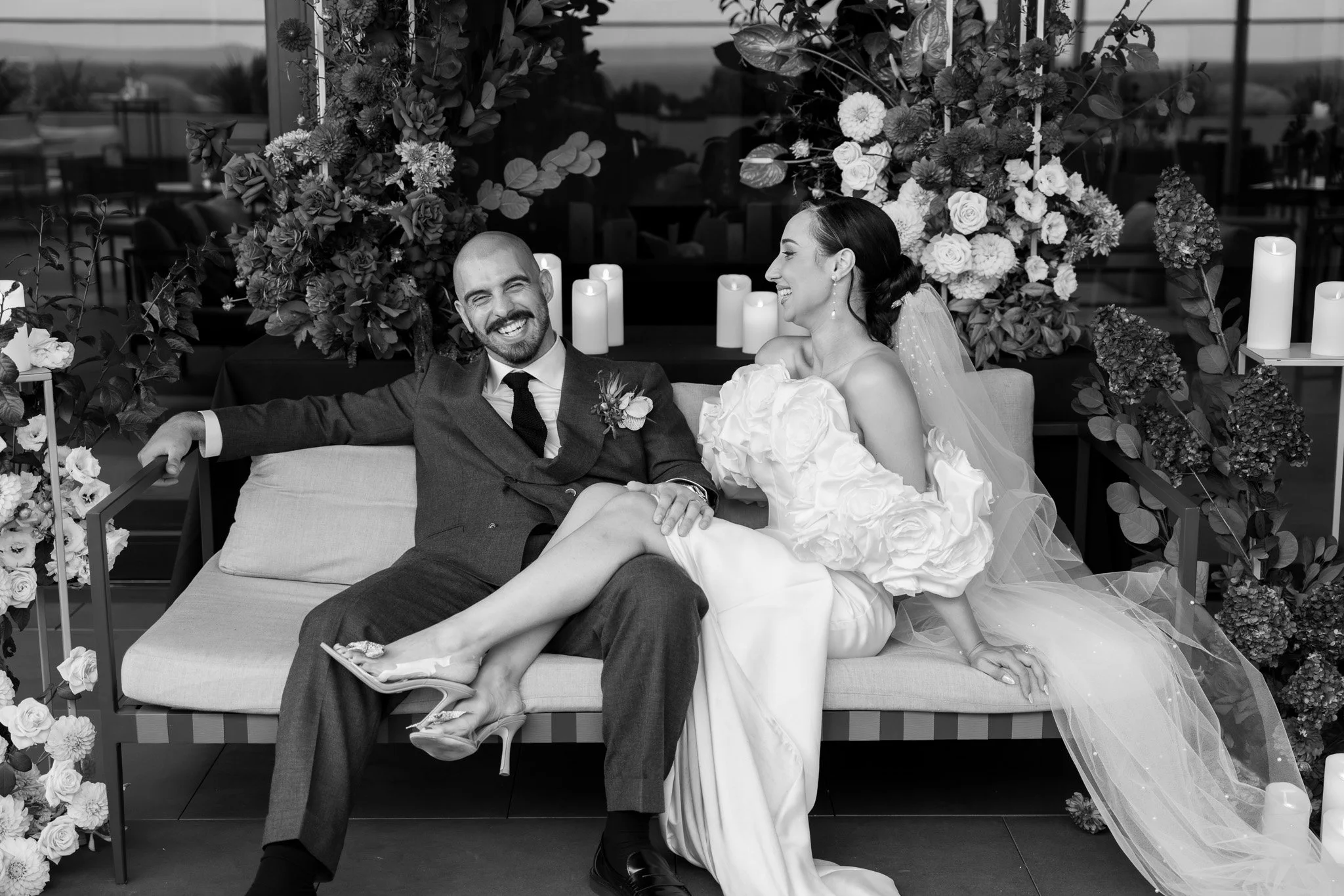 A smiling bride and groom sitting together on a bench, surrounded by floral decorations and candles, at their wedding celebration.