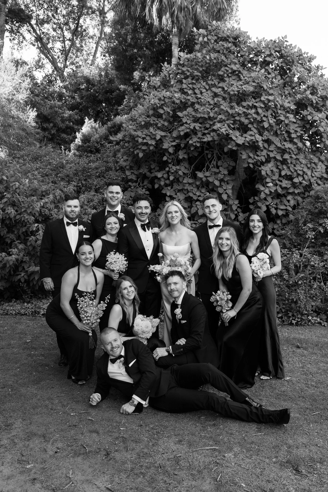 A wedding party with men in tuxedos and women in black dresses, several holding bouquets, gathered outdoors on grass in front of large trees and bushes, posing for a group photo.
