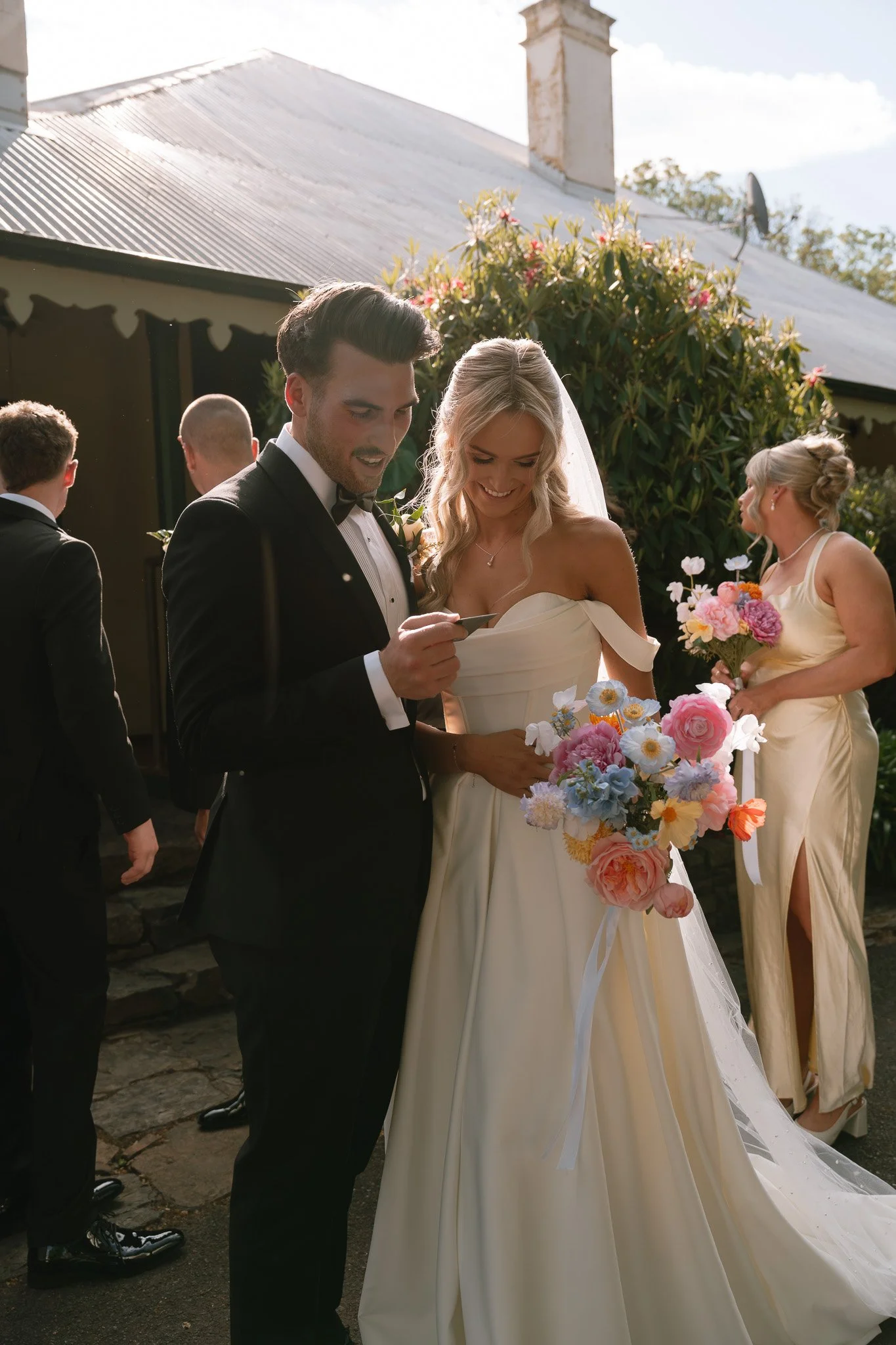 A bride and groom at their wedding, standing outdoors, looking at a piece of paper, with a bridesmaid holding a bouquet in the background.