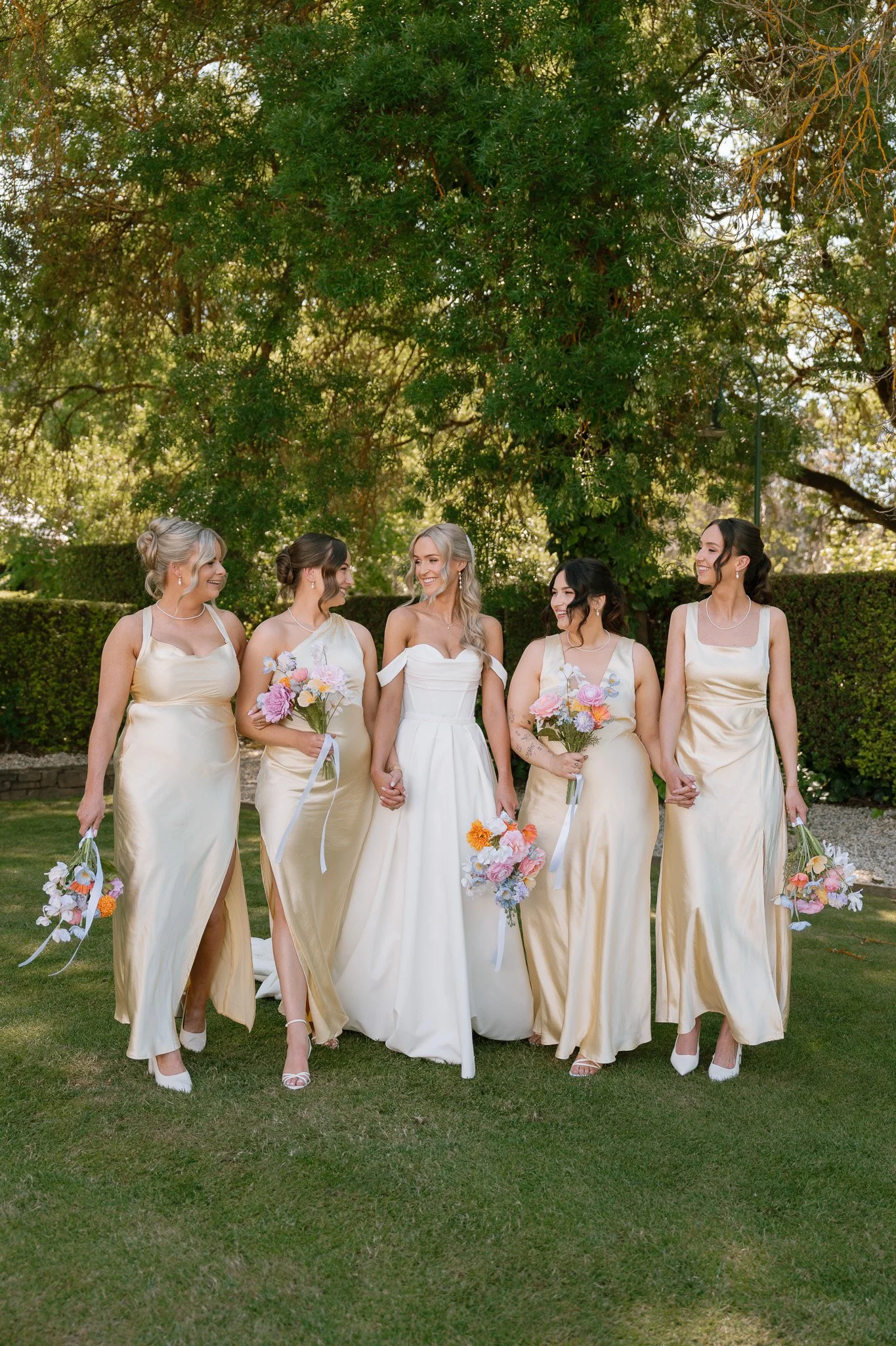 A bride and her four bridesmaids walking outdoors on a grassy area with trees in the background. The bride is in a white wedding gown, and the bridesmaids are in matching cream-colored dresses, all holding bouquets of pink, white, and orange flowers.