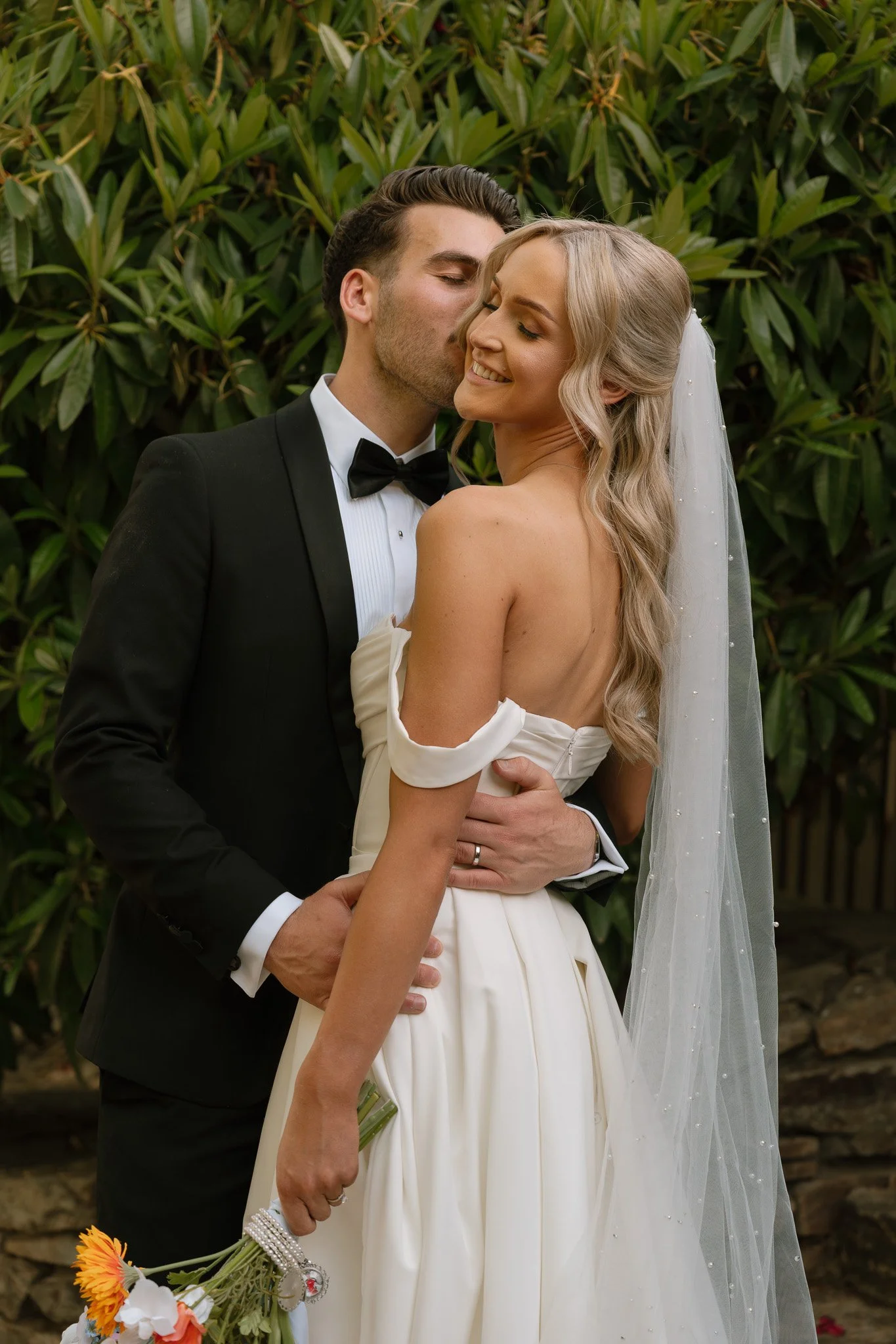 A newlywed couple sharing a romantic moment outdoors, with the groom kissing the bride on the cheek. The groom is in a black tuxedo with a bowtie, and the bride is wearing a white off-shoulder wedding gown with a veil. The bride is holding a bouquet 