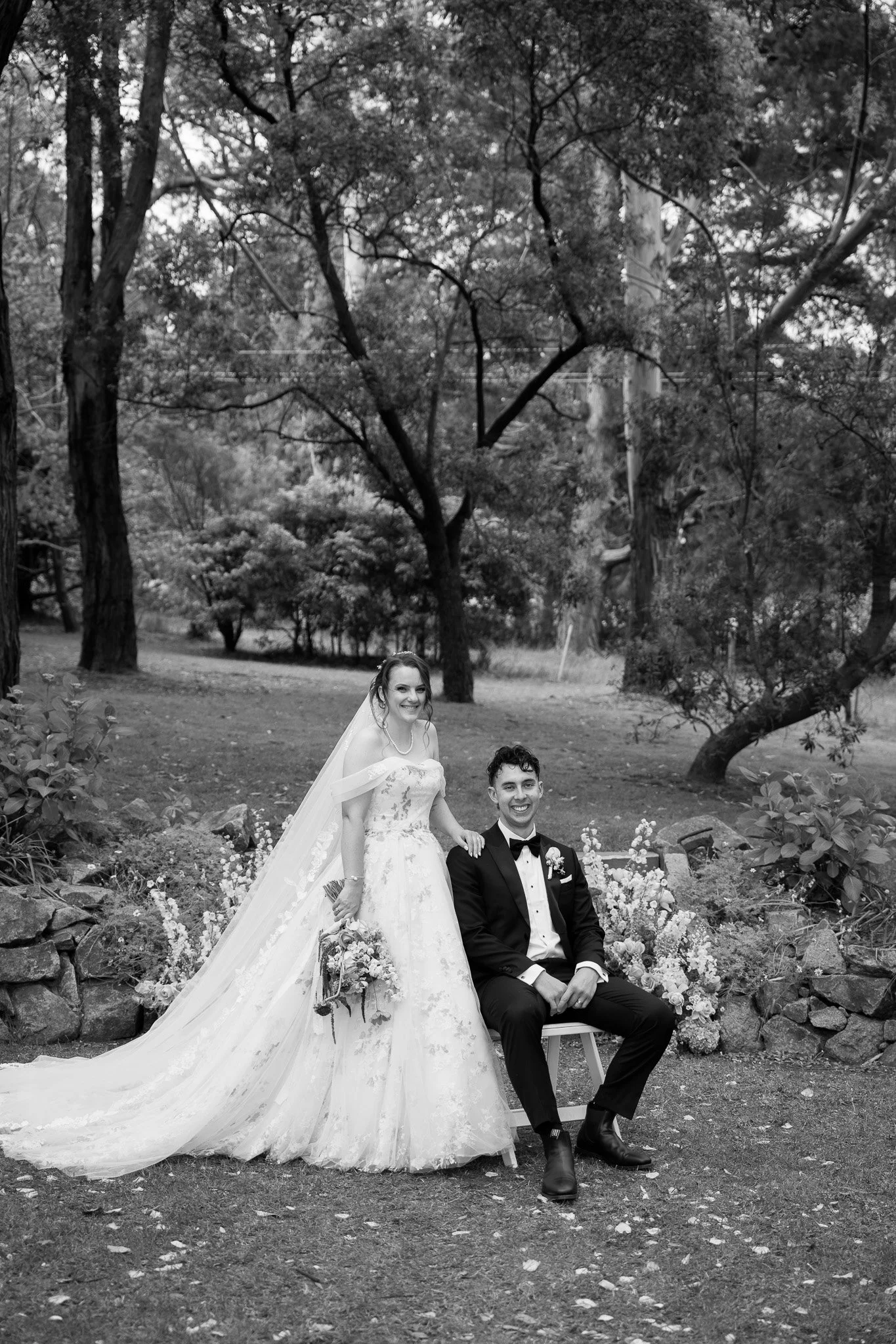 A wedding couple outdoors, with the bride standing and the groom sitting on a small stool, surrounded by trees and flowers, in a black and white photo.