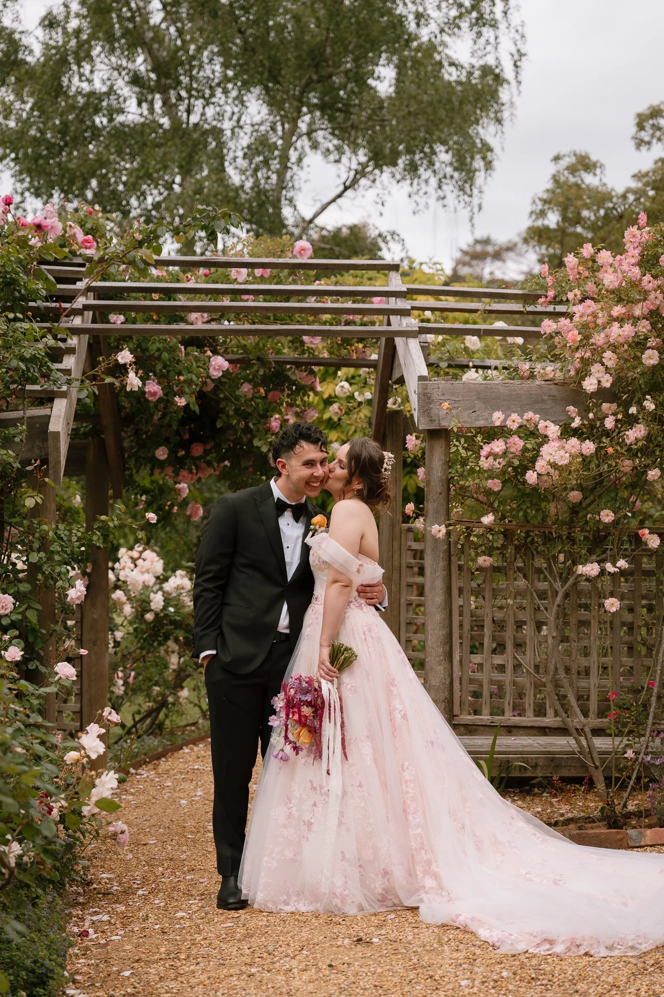 A bride and groom stand close together in a garden archway surrounded by pink roses. The bride, in a pink wedding gown, holds a bouquet, and the groom, in a black tuxedo, kisses her on the cheek.