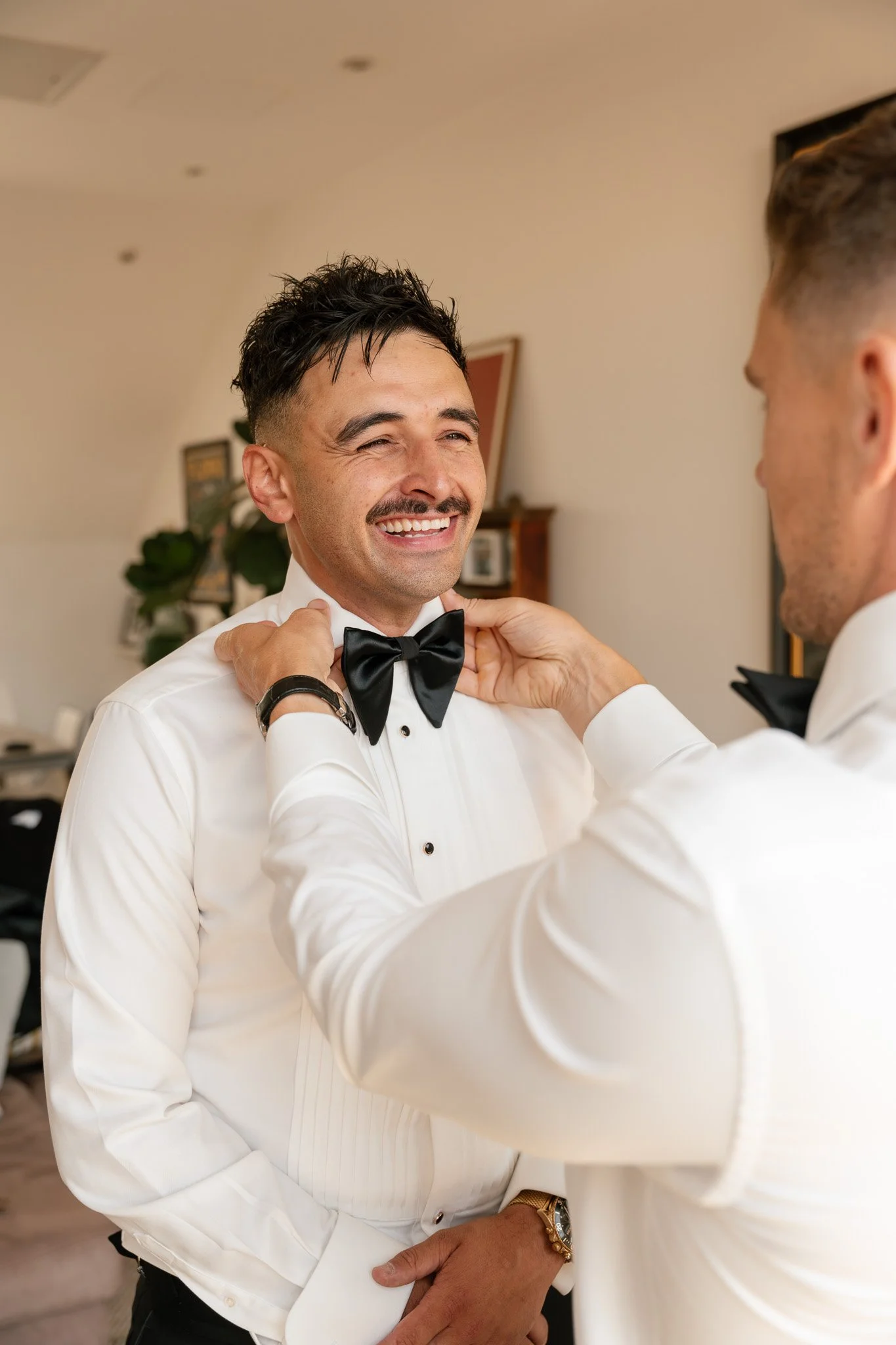 Two men in tuxedos preparing for a formal event, with one adjusting the other's bow tie while smiling.