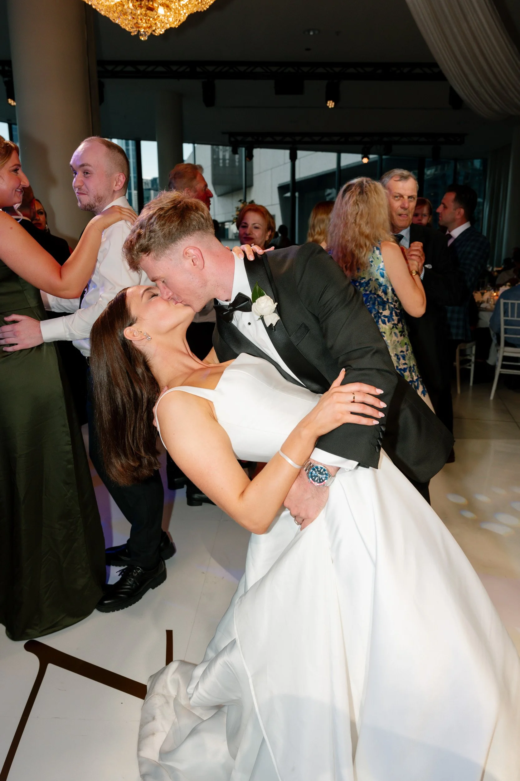 A bride and groom share a kiss during their wedding reception, surrounded by dancing guests at an elegant indoor venue with large windows and a chandelier.