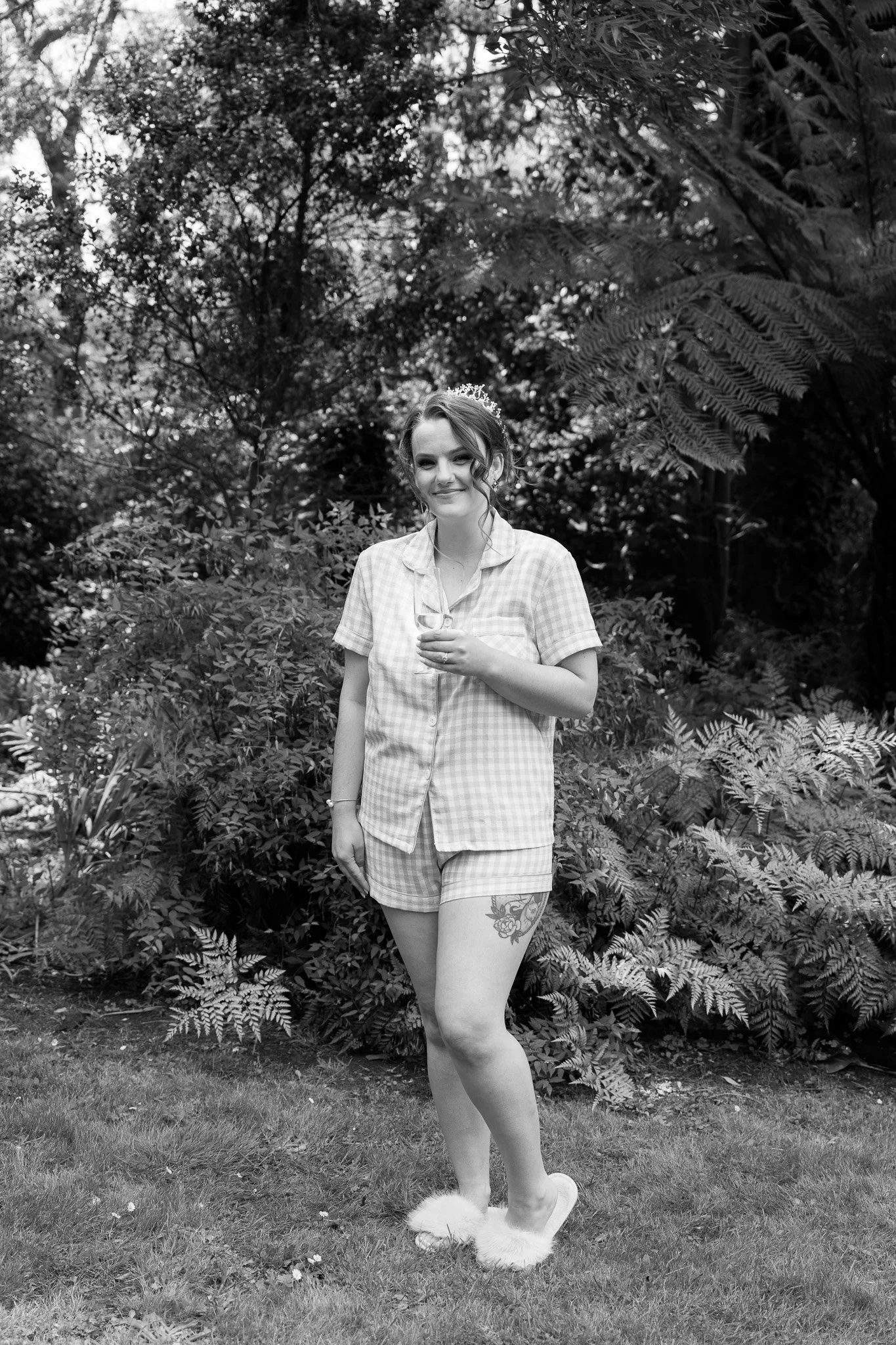 A young woman with a tattoo on her thigh standing on grass in pajamas and fuzzy slippers, holding a glass, in front of a garden.