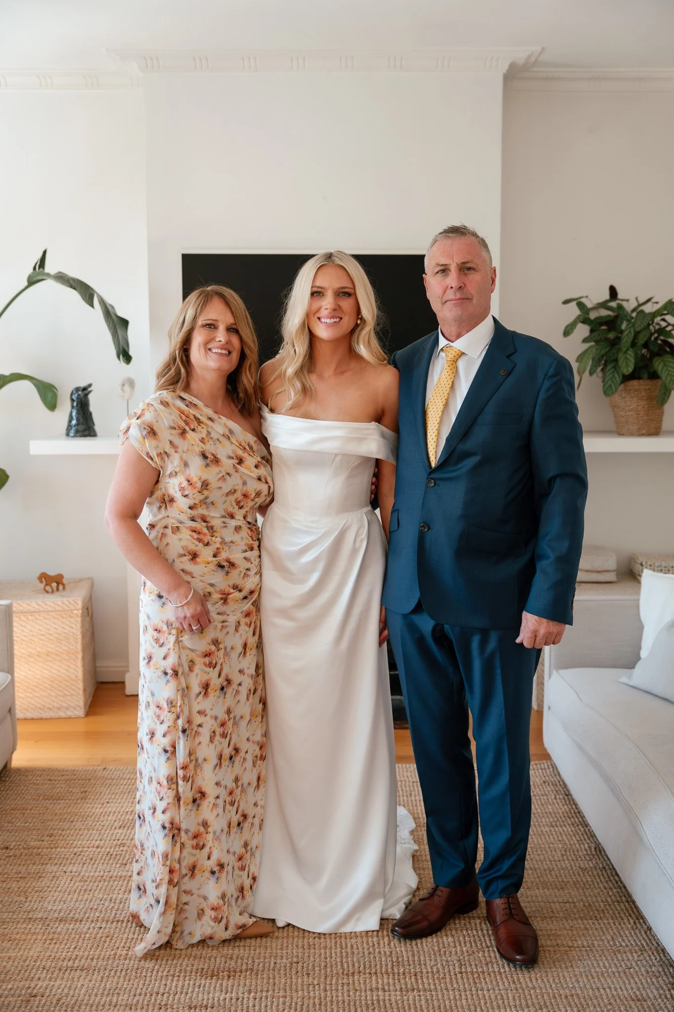 Three people standing in a living room, dressed in formal attire, smiling, with a black fireplace screen and potted plants in the background.