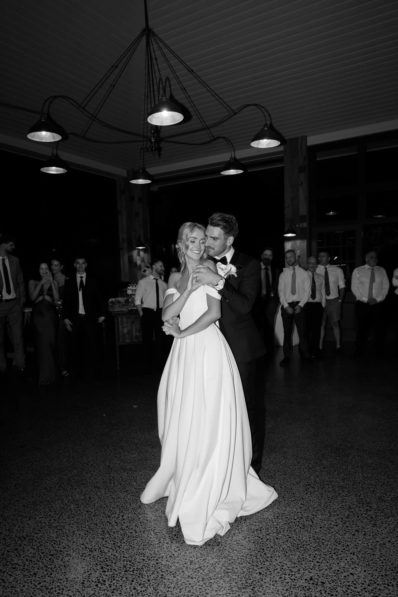 Black and white photo of a bride and groom dancing at their wedding reception, surrounded by guests.