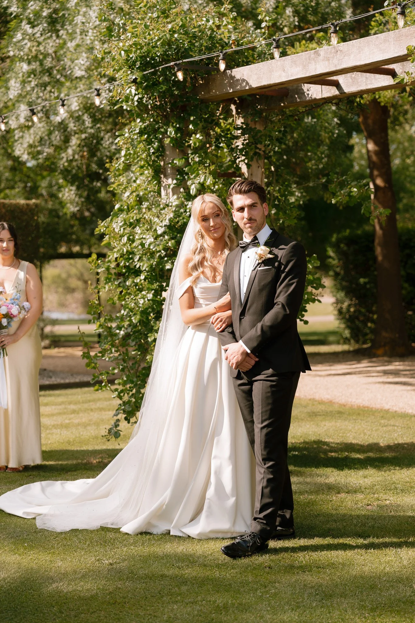 A bride and groom standing outdoors during their wedding ceremony. The bride is wearing a white wedding dress with a long train and veil, and the groom is dressed in a black tuxedo with a bow tie. A bridesmaid in a cream dress holding a bouquet stand