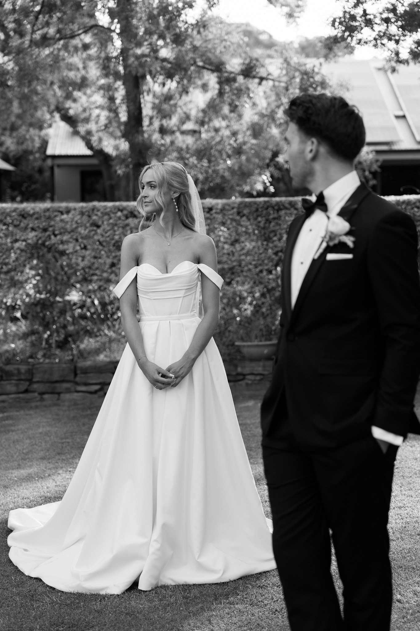 Black and white photo of a bride and groom outdoors, with the bride in a strapless wedding gown and the groom in a tuxedo, standing on a lawn with trees and a house in the background.