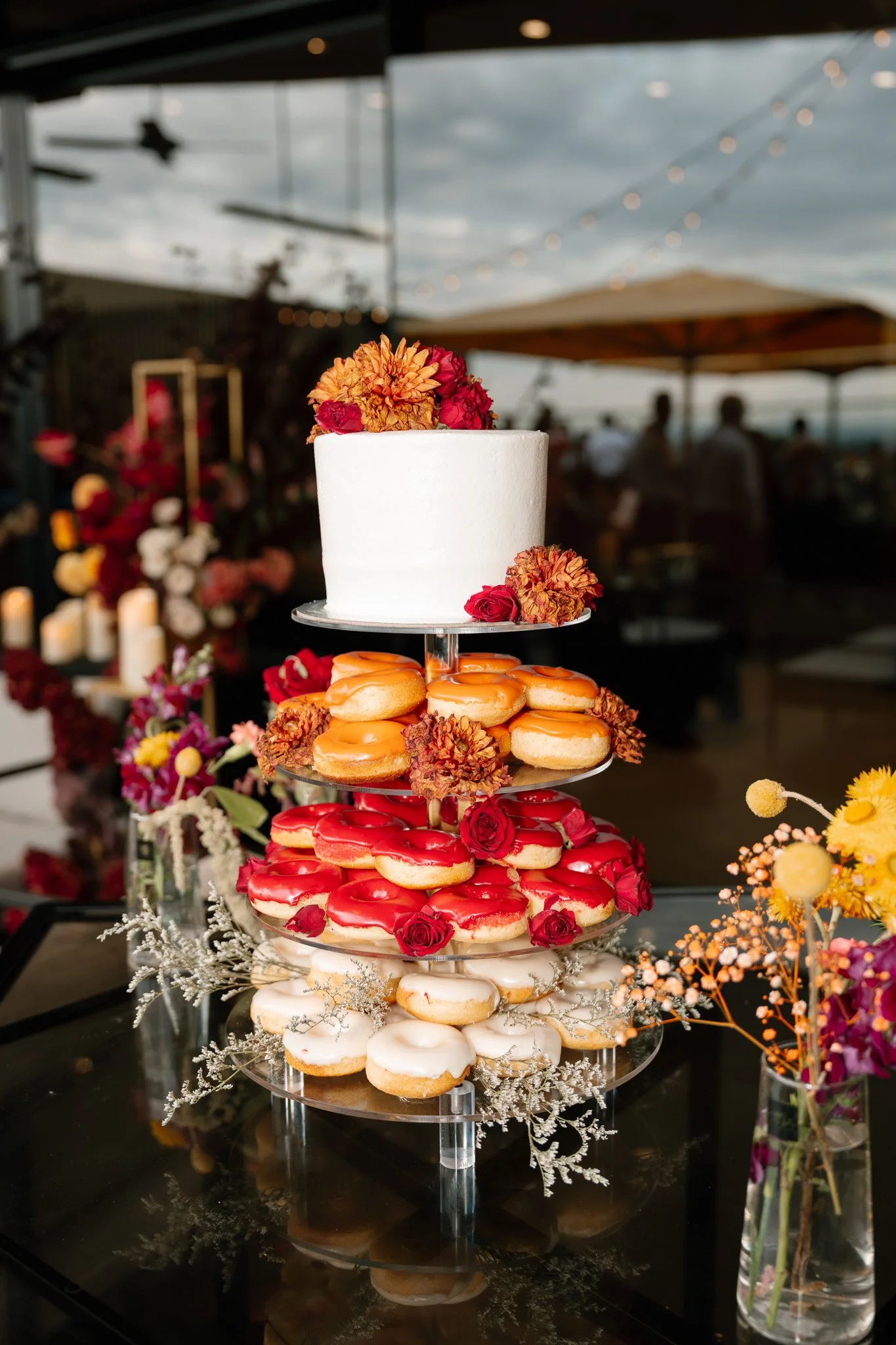A four-tiered dessert stand with a white cake on top, decorated with orange and red flowers. The lower tiers hold decorated donuts with red and white icing, accented with roses and flowers. The setup is outdoors, surrounded by flowers and with a clou