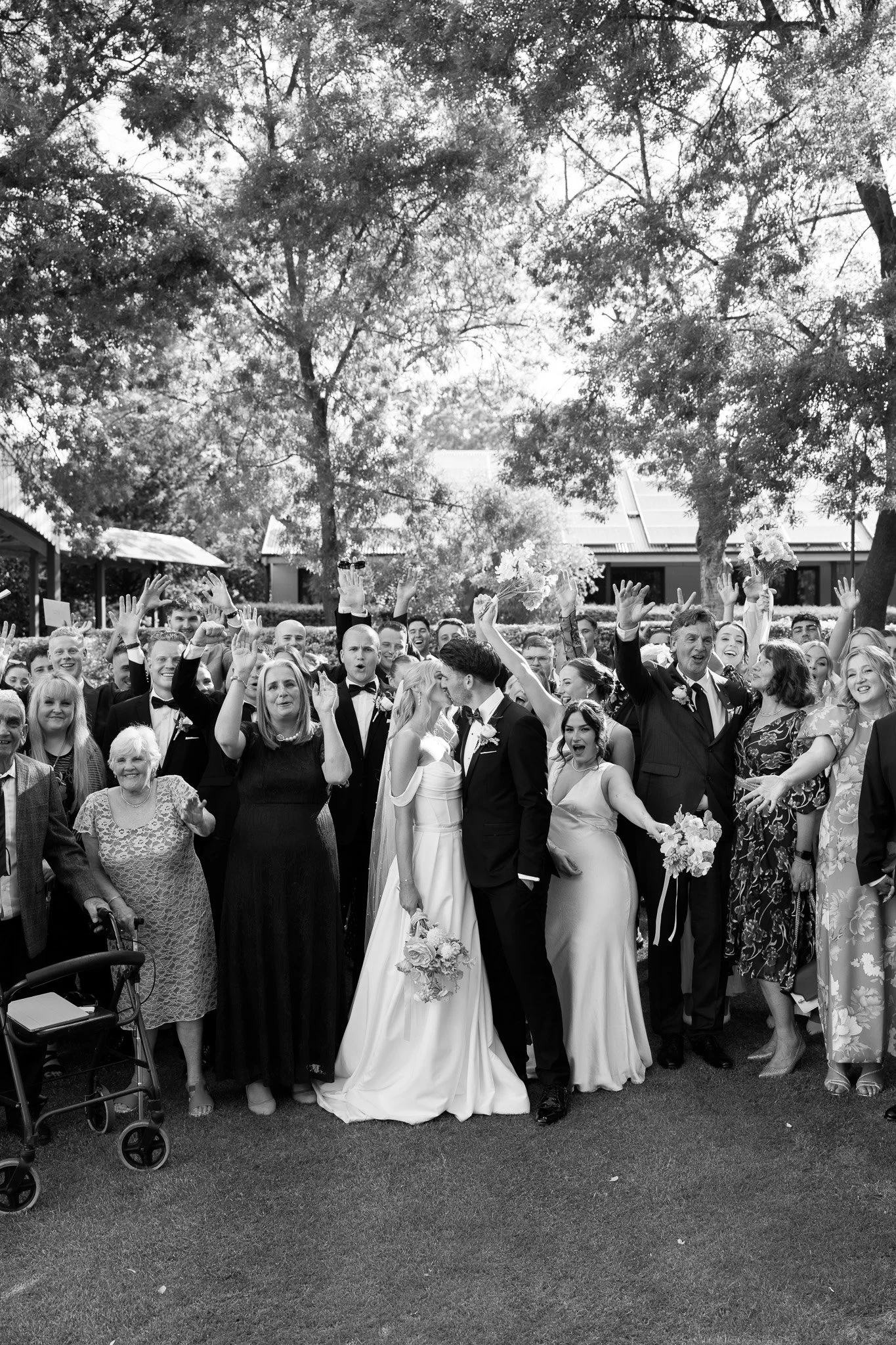 Black and white photo of a wedding celebration with a group of people standing outdoors, celebrating the newlywed couple who are kissing in the center. The crowd is smiling, raising hands, and holding flowers, surrounded by trees and buildings in the