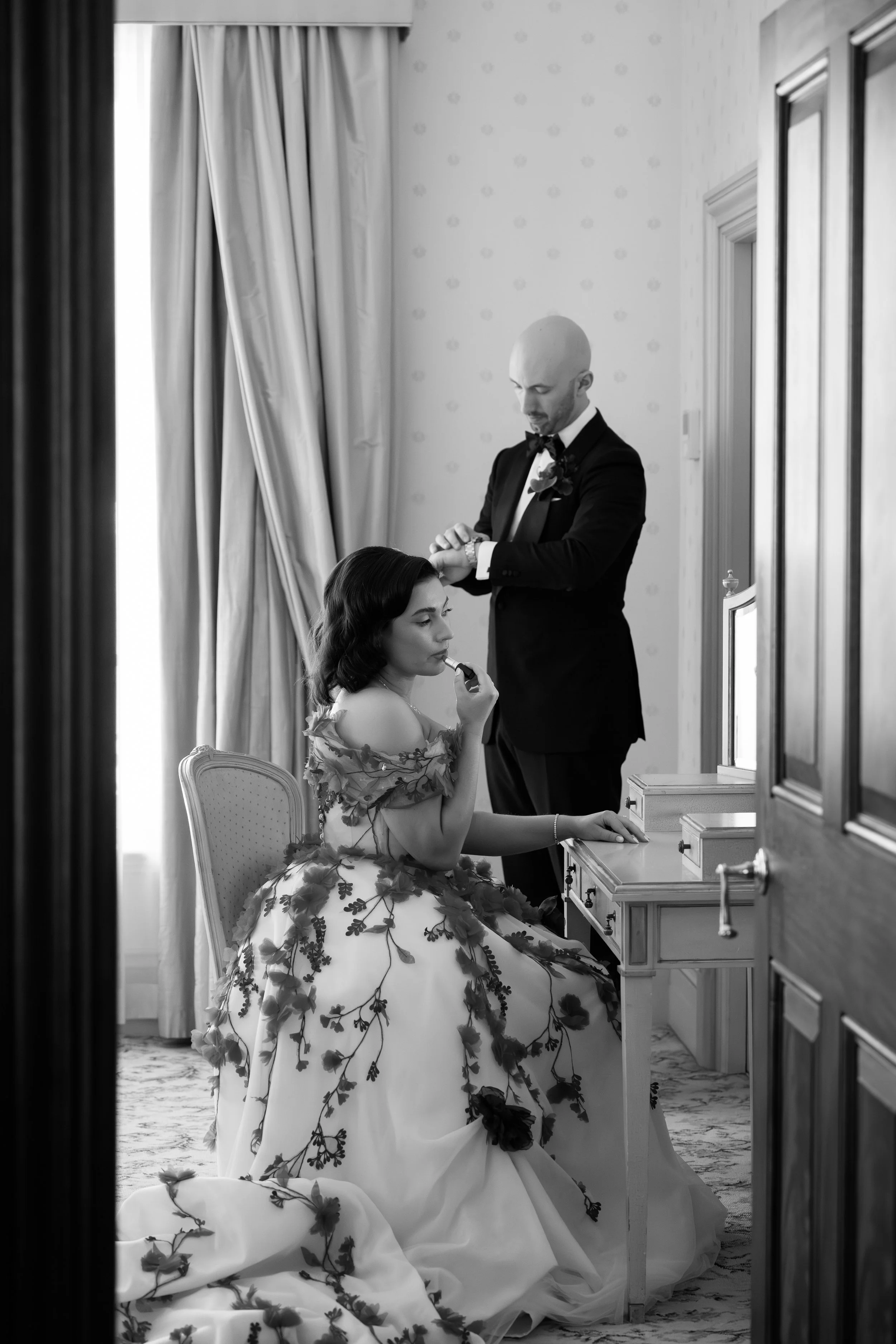 A man in a tuxedo assists a woman in a wedding dress with makeup, sitting at a vanity. The scene is in a well-lit room with curtains.