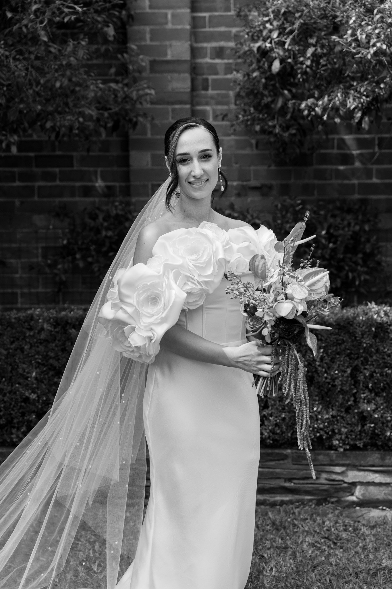 Black and white photo of a smiling bride in a wedding dress holding a bouquet of flowers, standing outdoors in front of a brick wall with bushes.