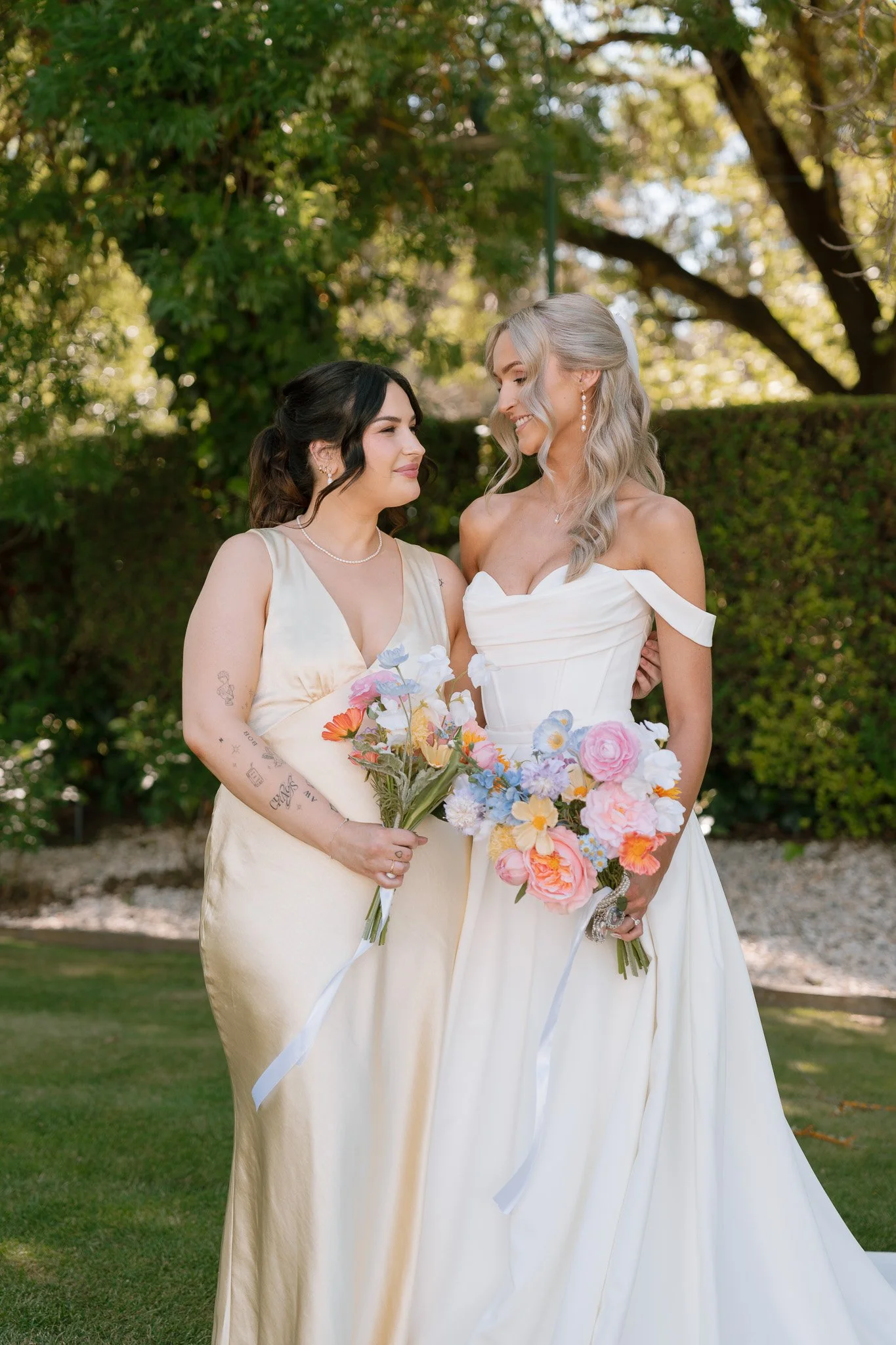 Two women in wedding dresses standing outdoors, smiling at each other, holding bouquets of flowers with pink, purple, and white blossoms against a green background.