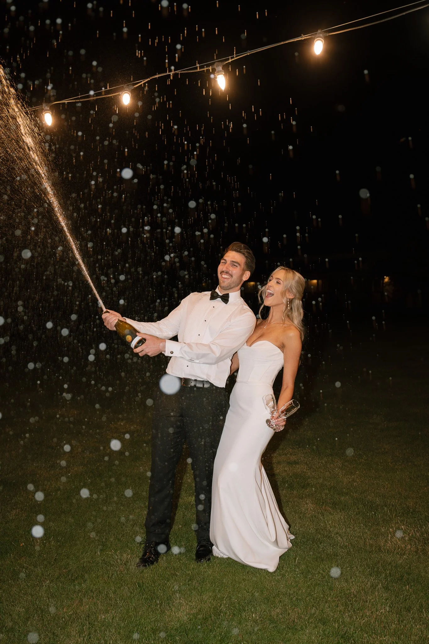 Happy bride and groom celebrating at night, spraying champagne, with string lights above, on a grassy outdoor area.