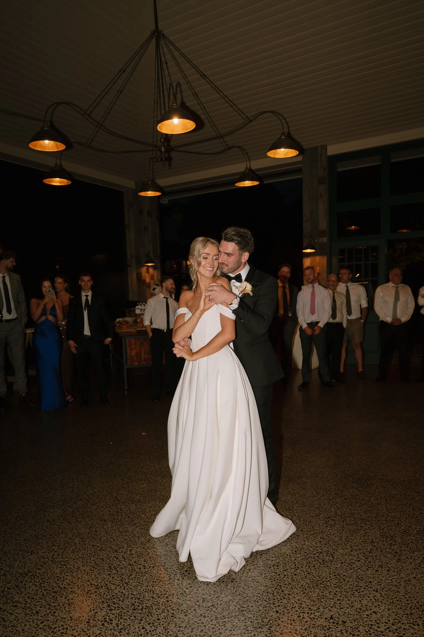 A bride and groom share their first dance at their wedding reception, surrounded by guests in a dimly lit venue.