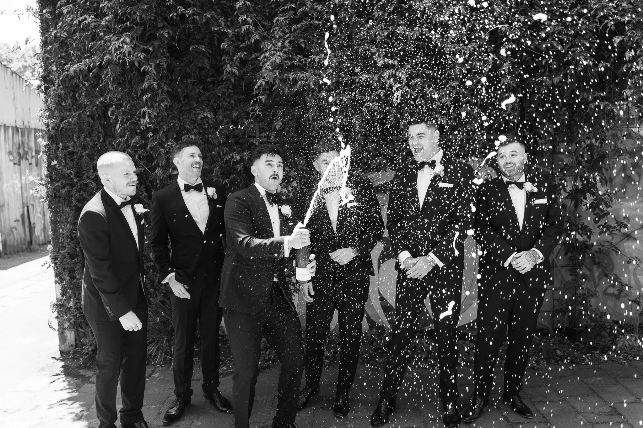 Group of men in tuxedos celebrating with a champagne pop and confetti outdoors.