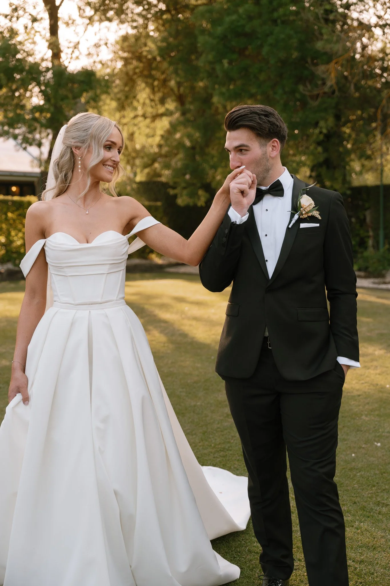 A bride and groom standing outside during sunset, with the bride touching the groom's nose, both smiling. The bride is wearing a white off-shoulder wedding gown and the groom is in a black tuxedo with a bow tie.