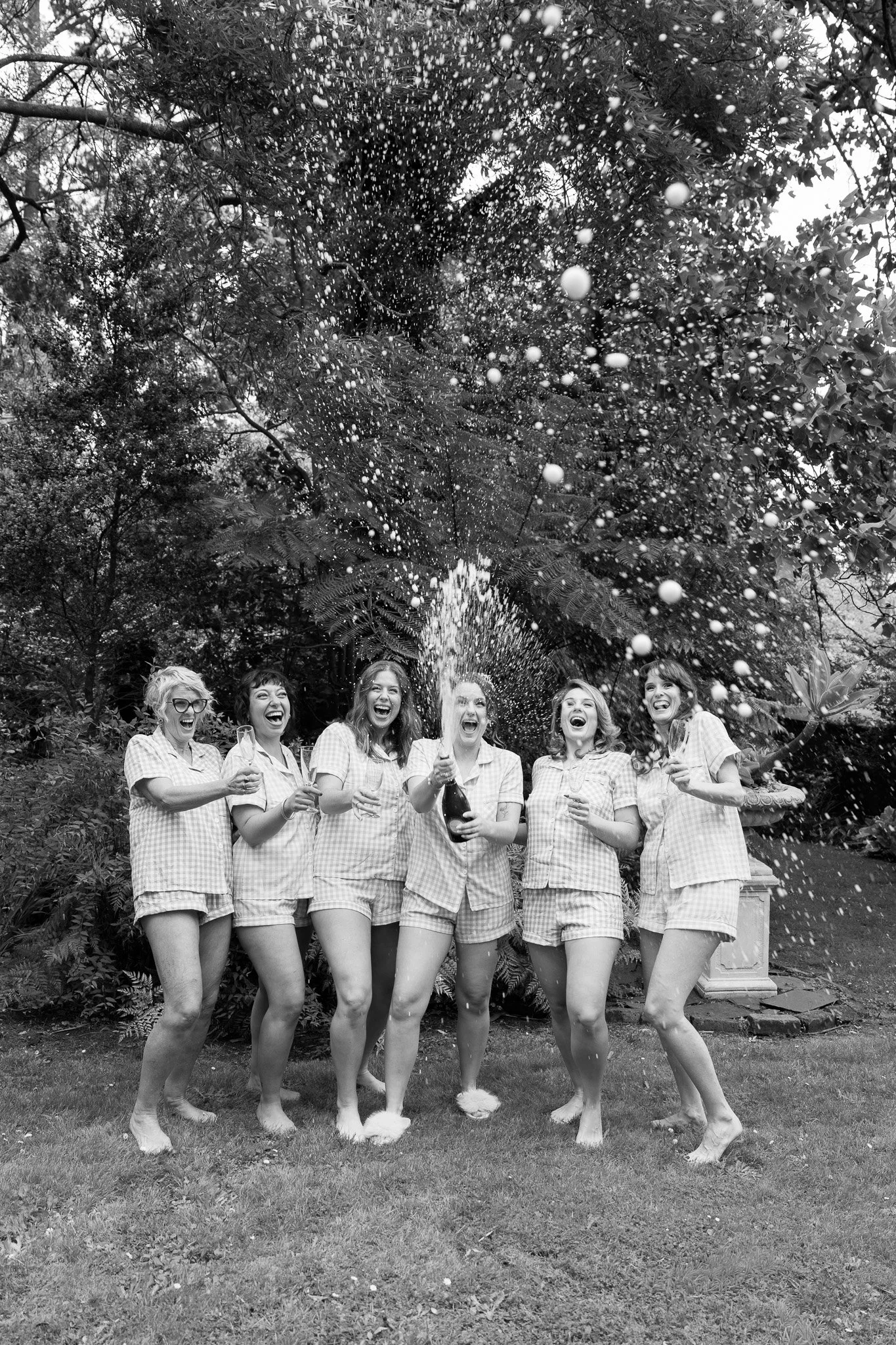 Seven women in pajamas celebrating outdoors, popping a champagne bottle, with spray of champagne in air, trees in background, black and white photo.