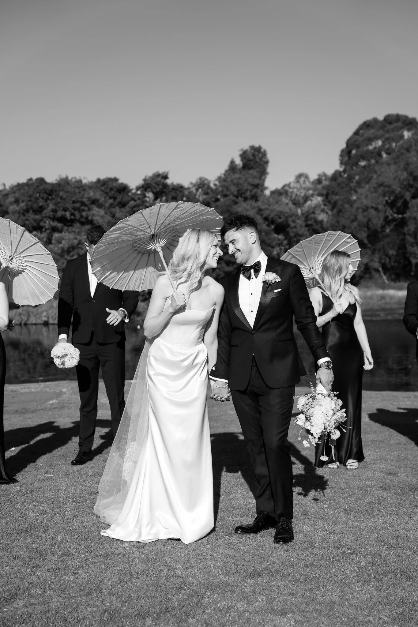 A black and white photo of a wedding scene outdoors. A bride and groom hold hands and lean towards each other, smiling. The bride is in a strapless wedding gown with a long veil, and the groom is in a tuxedo. Several people, some holding umbrellas, s