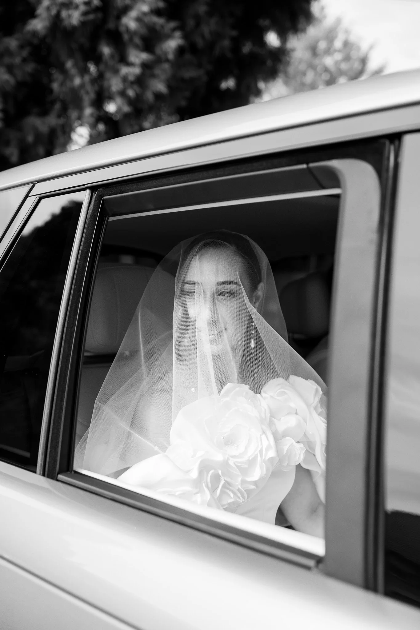 A bride in a wedding dress and veil sitting inside a car, smiling and looking out the window.