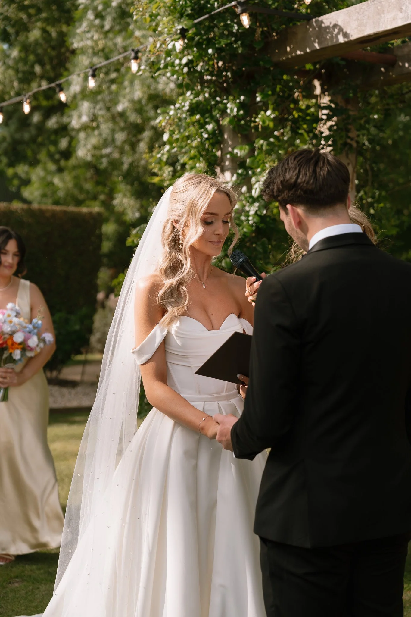 Bride and groom exchange vows outdoors during wedding ceremony, with bridesmaid holding bouquet in background.